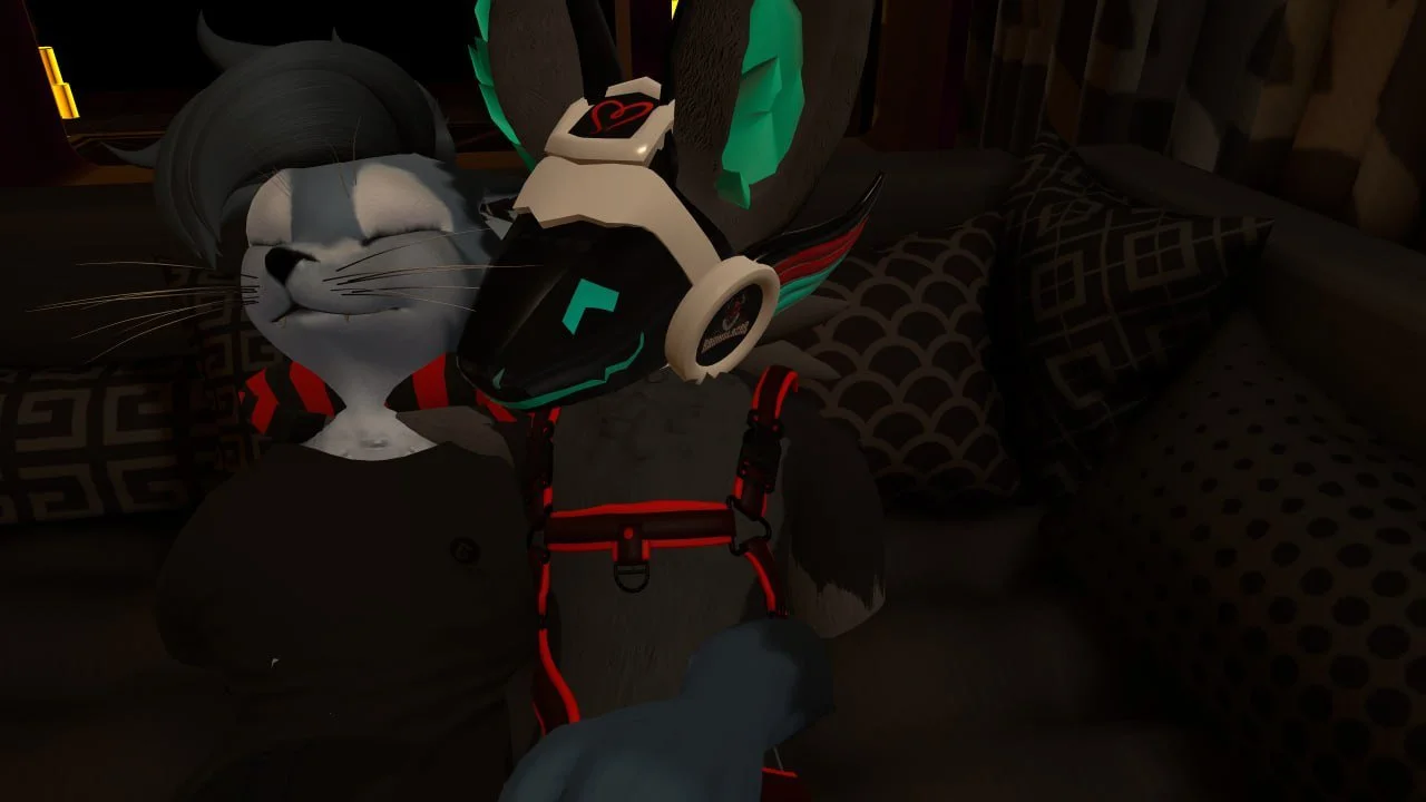 A computer-generated anthropomorphic cat character with gray and white fur, wearing a black shirt, red harness, large white headphones, and gaming gear, standing in a dimly lit room with patterned pillows on a sofa in the background.