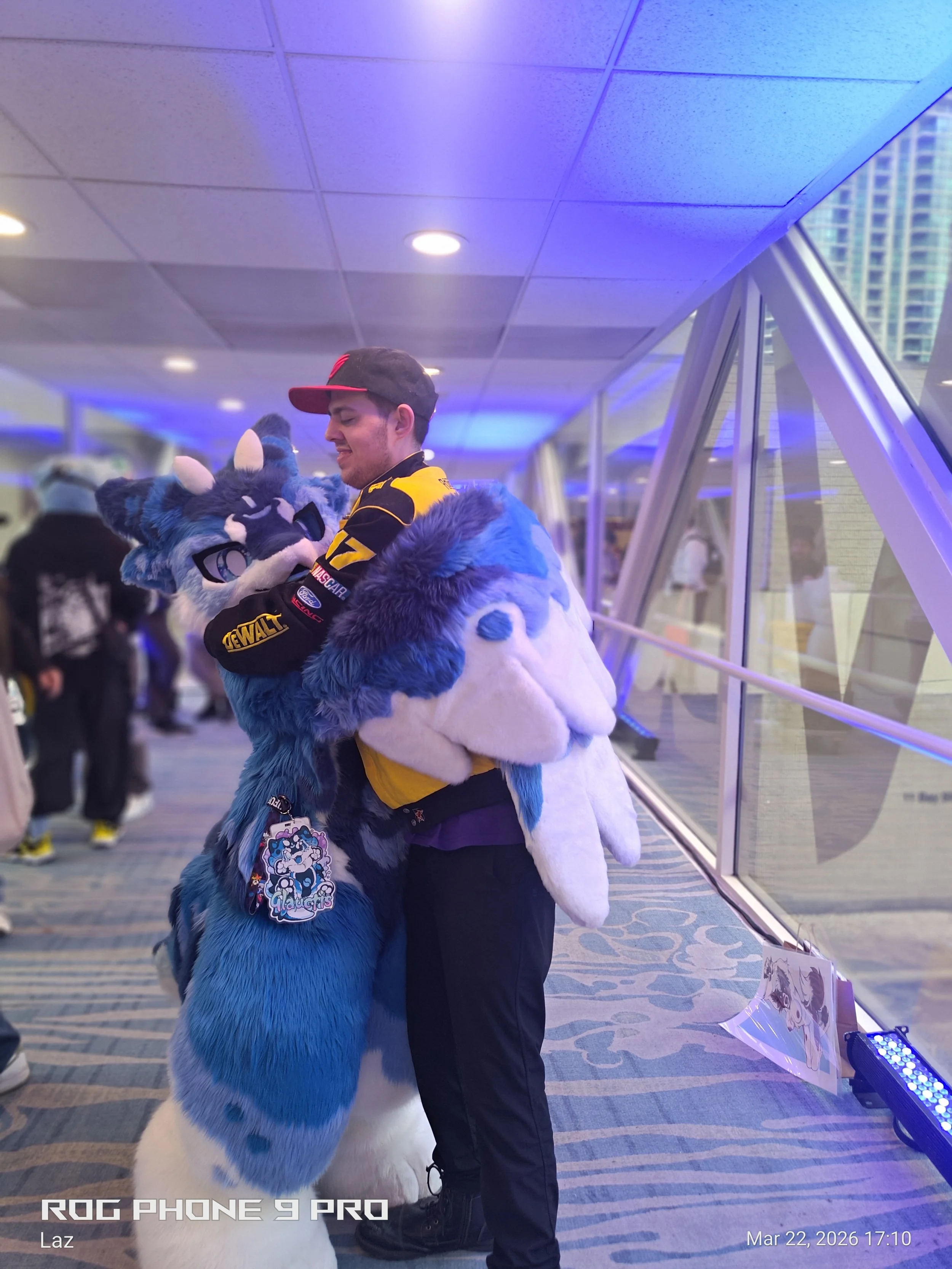A person in a black racing suit with yellow accents, posing with a person in a wolf mascot costume in an indoor setting with a large window and city buildings outside. The mascot is blue and white with a tag hanging from its collar. The timestamp on 