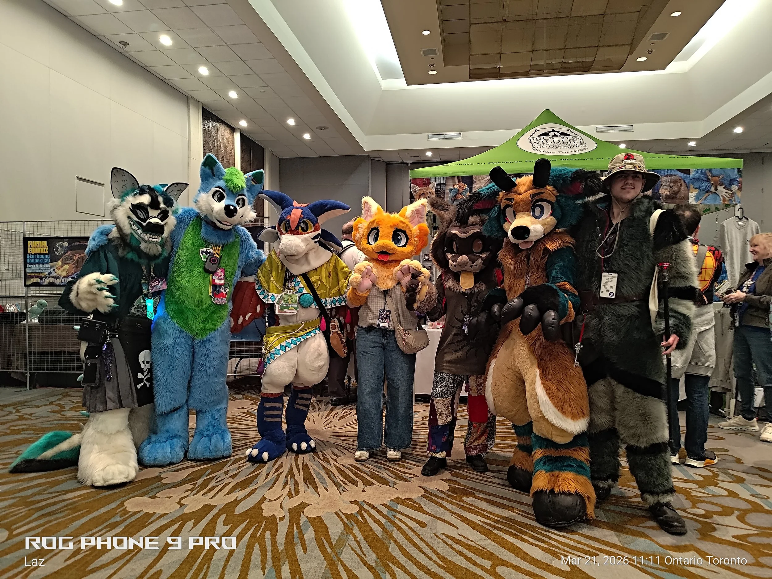 Group of people dressed in various fursuits and costumes at a convention, standing in front of a green tent with a Yukon Wildlife logo.
