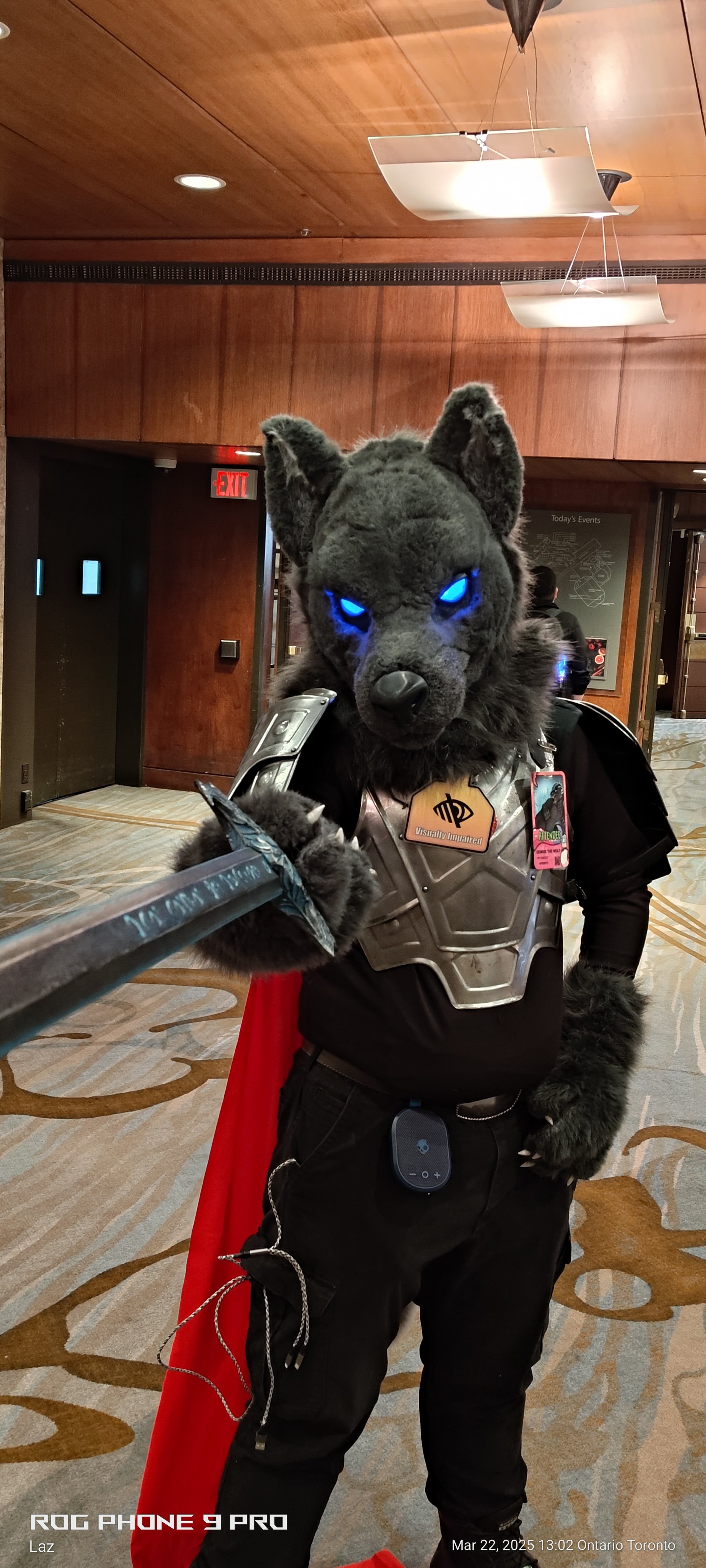 Person dressed in a wolf-like costume with glowing blue eyes, wearing a costume armor, taking a selfie with a sword, in an indoor setting with wooden walls and a patterned carpet.