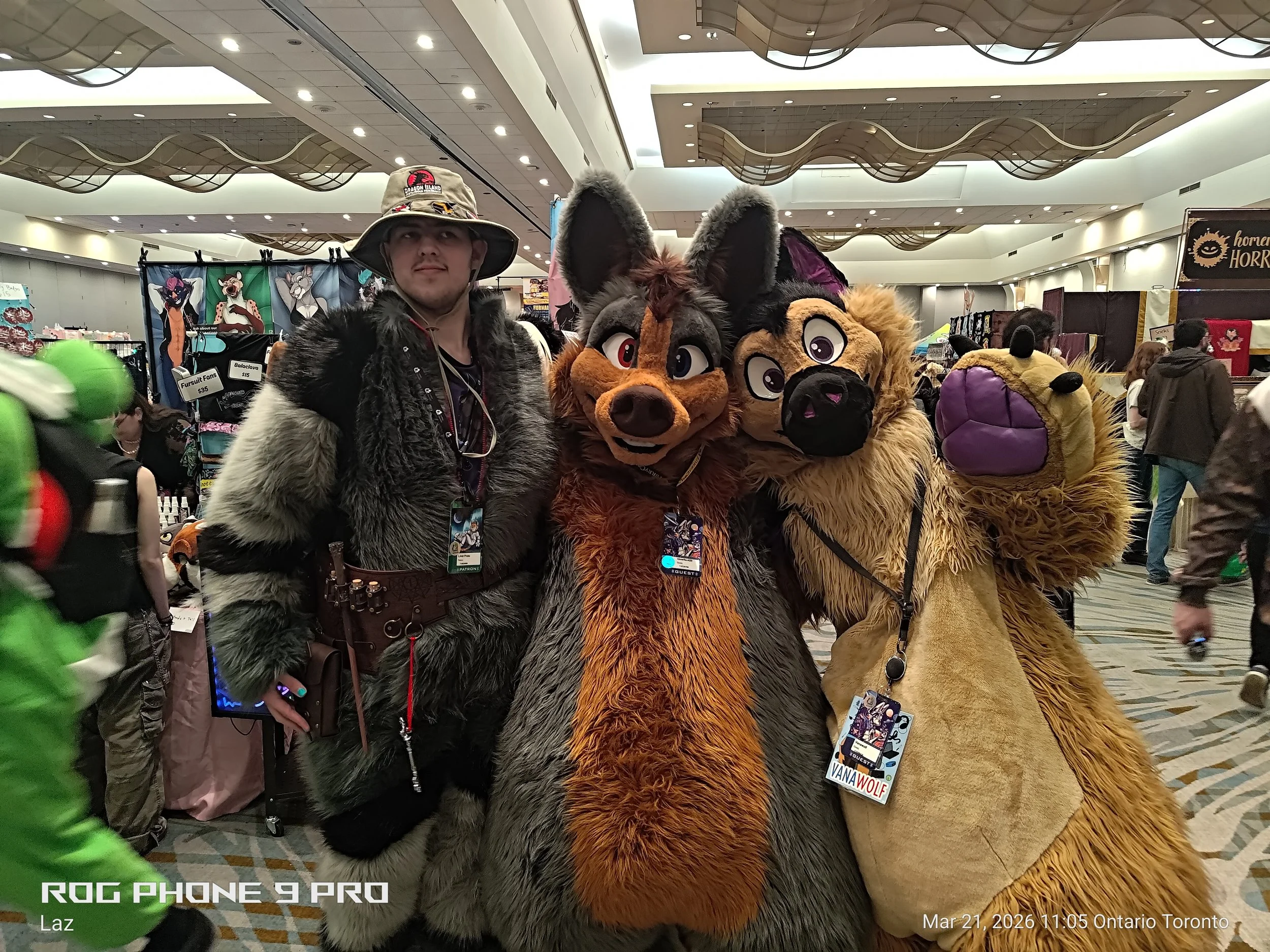 People dressed in colorful furry animal costumes at a convention, with vendor booths and other attendees in the background.