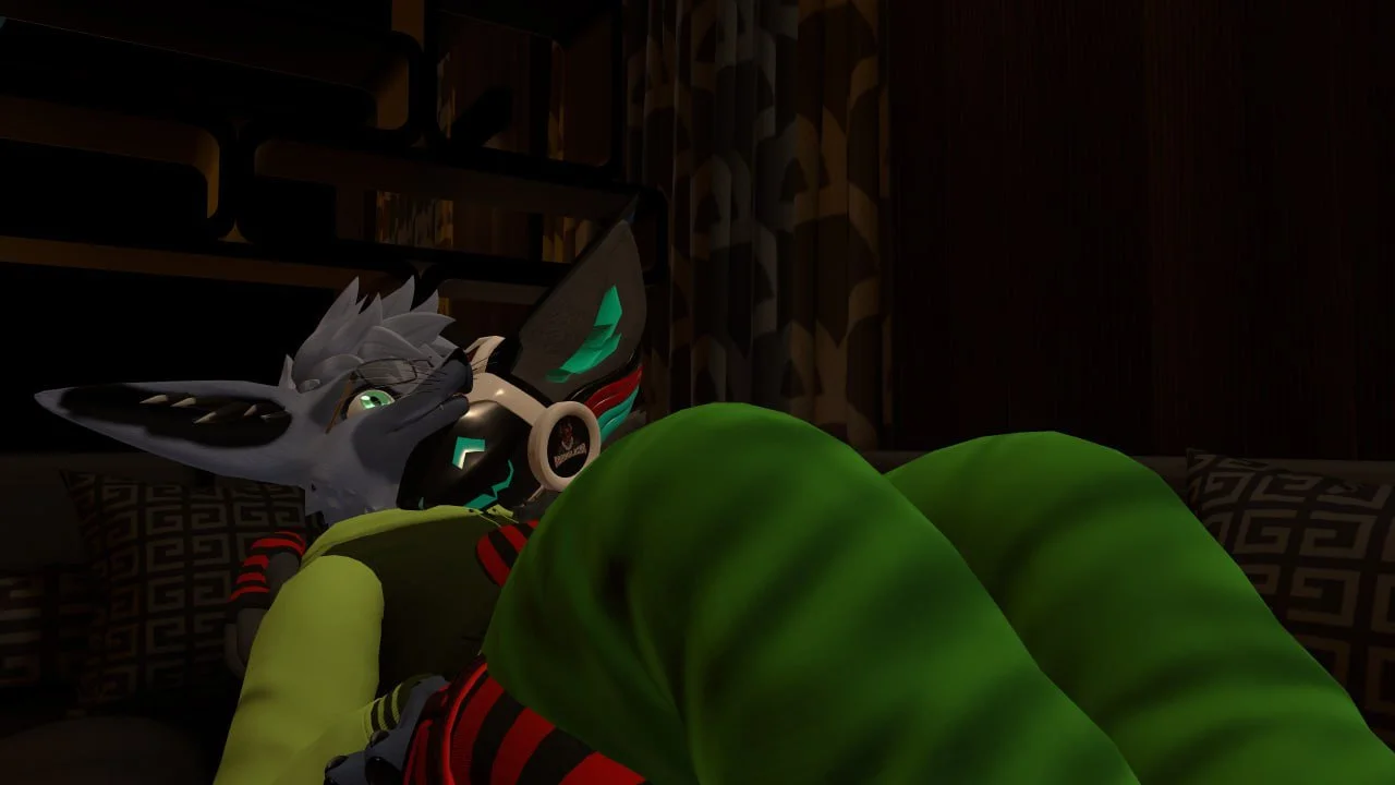A humanoid fox character with gray fur, green eyes, and glasses, lying on a bed in a dark room, holding a gaming controller and wearing colorful pajamas.