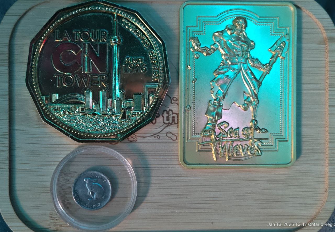 A collection of three metallic items: a large, octagonal coin decorated with a city skyline and the text 'La Tour CN Tower Since 1976,' a rectangular holographic item featuring a pirate holding a sword and shield with text at the bottom, and a small 