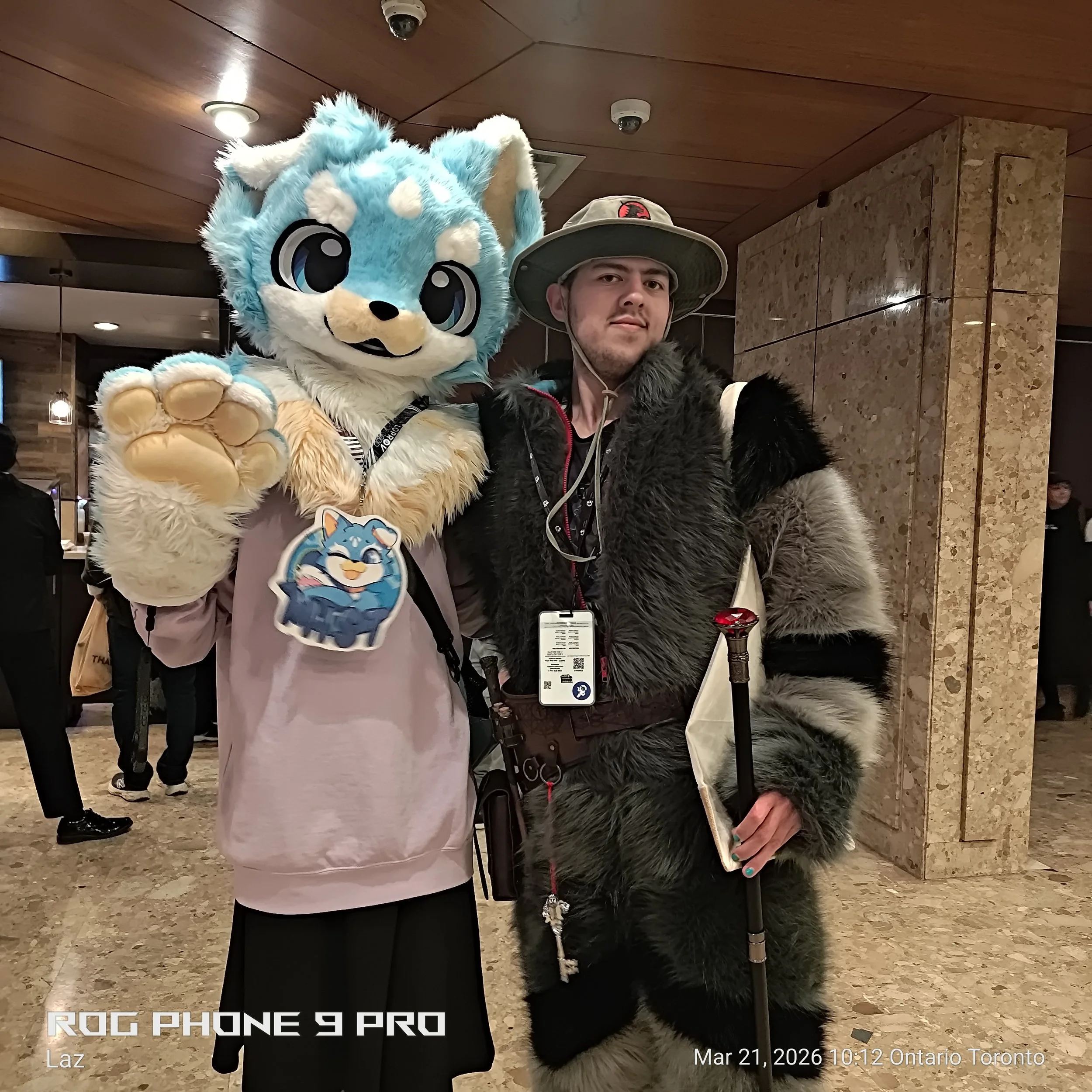 A man wearing a large hat with a Jurassic Park logo and a thick fur coat standing next to a person in a colorful, furry animal costume with large eyes and paw details, inside a building with wooden ceiling panels and marble or tiled walls.