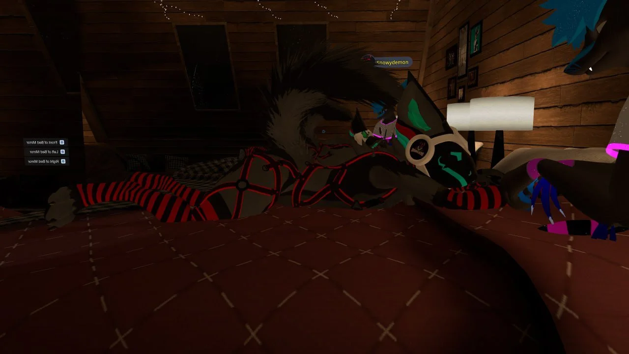 A digital avatar of a humanoid character with a black and teal face, wearing a striped red and black bodysuit, lying on a bed in a dimly lit wooden room. The room features a nightstand with a lamp, framed pictures on the wall, and a poster of a shark