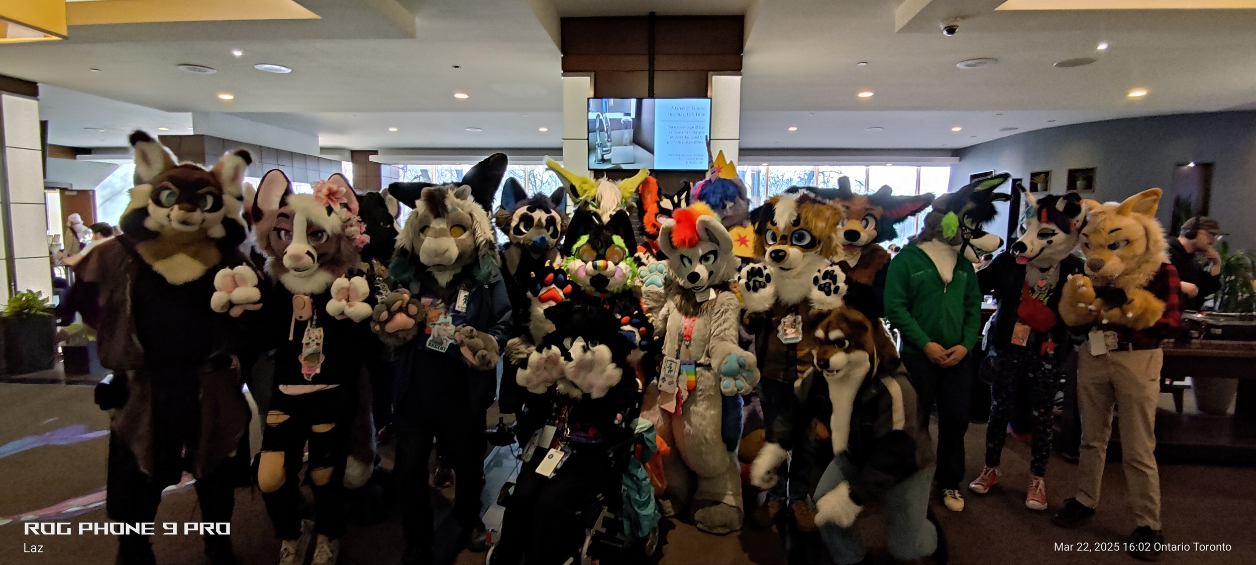 Group of people in fursuits at a convention, standing indoors, with a large monitor and windows in the background.