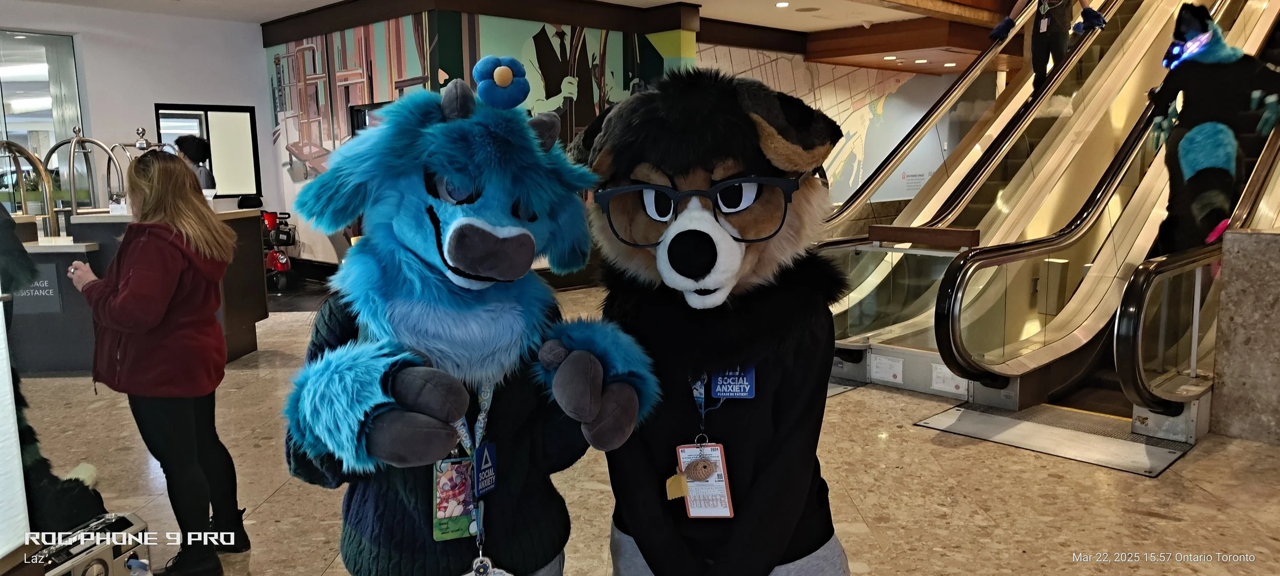Two people in furry animal costumes posing for a photo at an indoor location, with escalators and other people in the background.