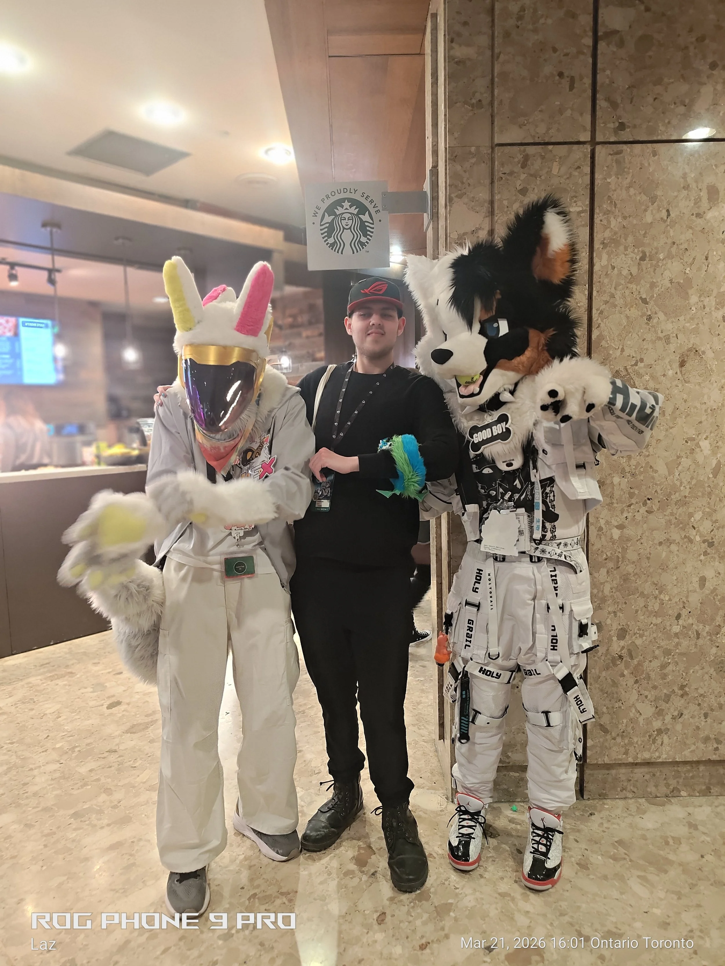 A person dressed in a bunny costume, a person dressed as a dog, and a person in black clothing standing together in a public indoor space with a Starbucks sign in the background.