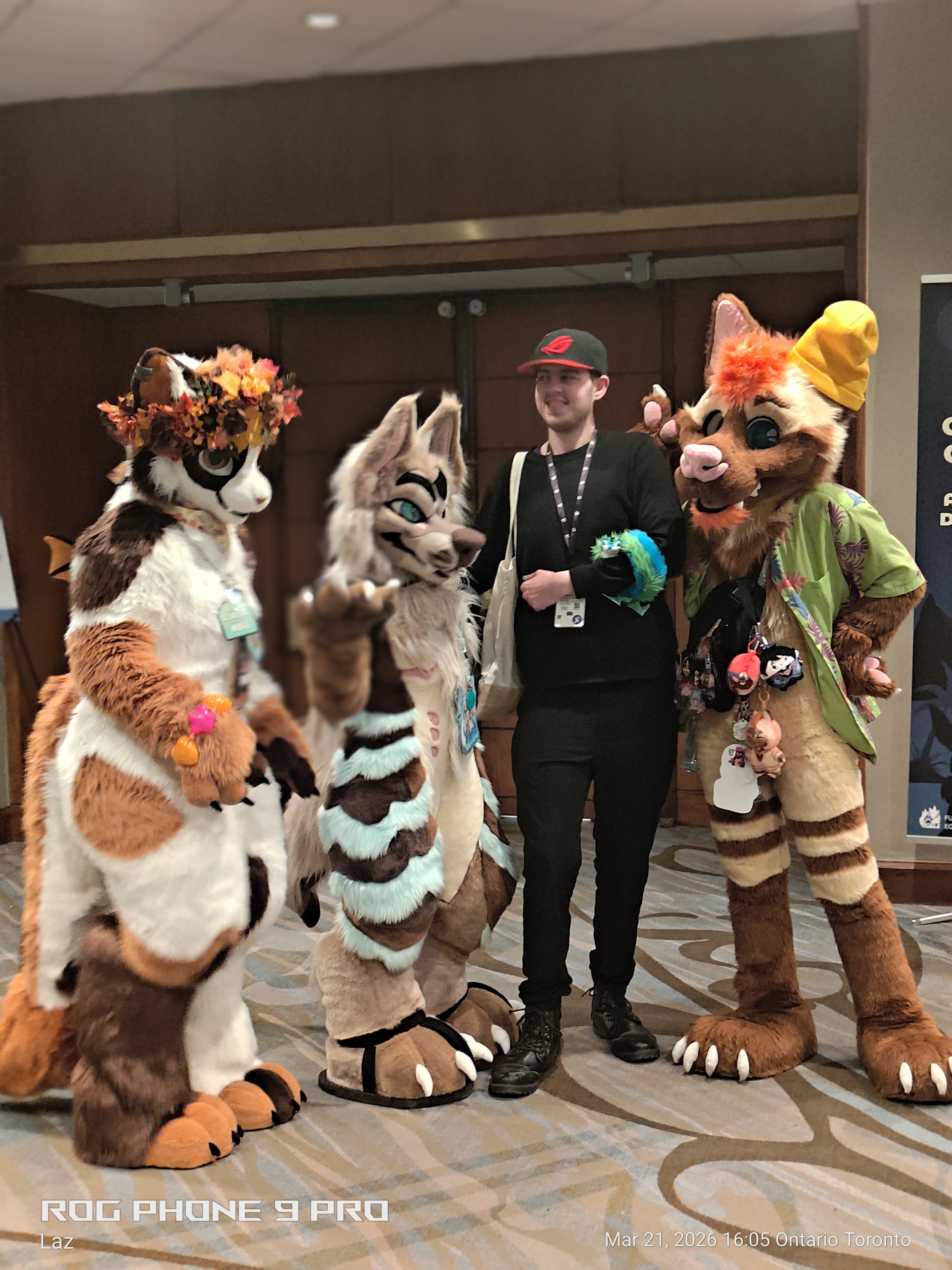 A group of people dressed in animal costumes posing for a photo at an indoor event, with a man in casual attire standing between two large anthropomorphic animal characters.
