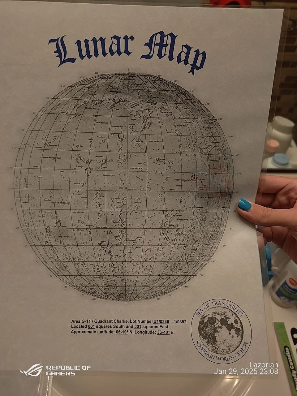 A person holding a lunar map with the title "Lunar Map" in blue Gothic font at the top. The map is a detailed illustration of the moon's surface with labeled features and grid lines. There is a stamp in the bottom right corner that reads "Sea of Tran