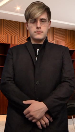 Young man in a black blazer and glasses standing indoors in front of wooden wall panels.