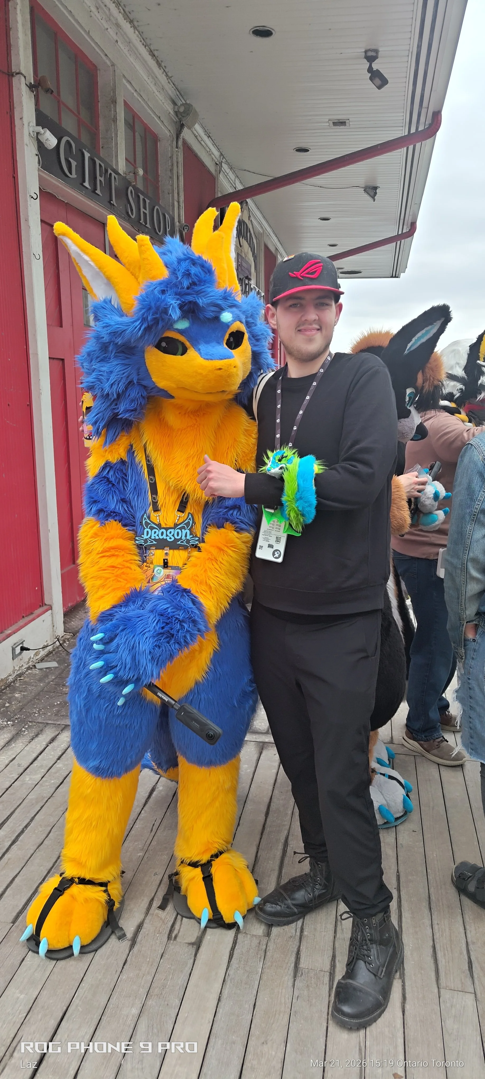 A person dressed in a colorful dragon costume standing beside a man in black clothing at a costume event outside a gift shop, with other costumed characters and people in the background.