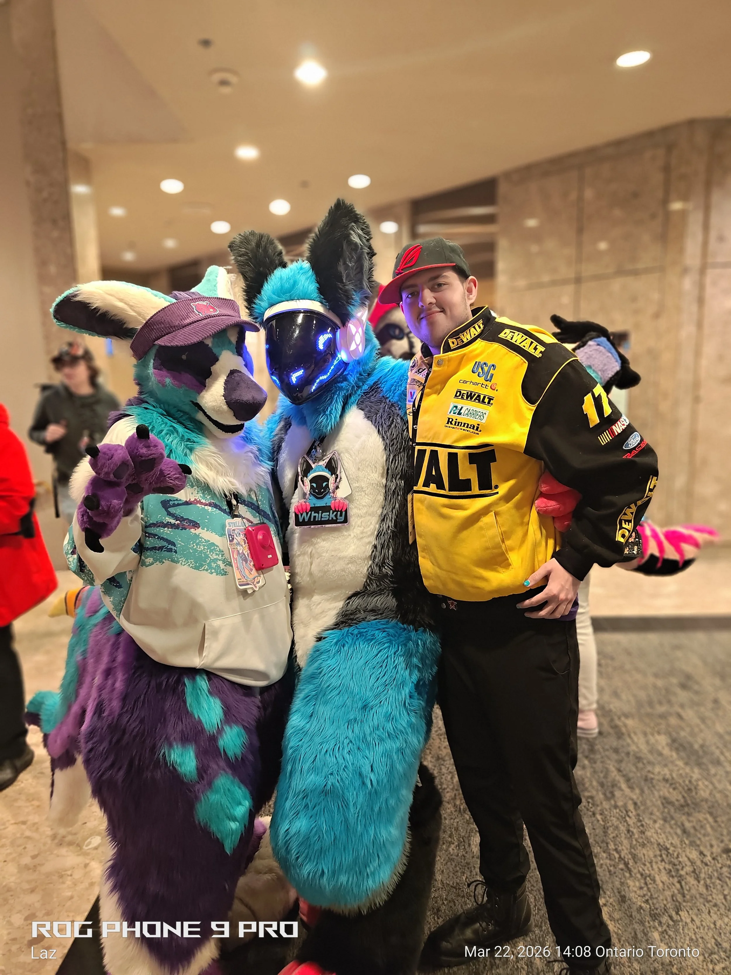 A man wearing a yellow racing jacket with sponsor patches, black pants, and a cap, standing indoors with two people in colorful furry animal costumes featuring blue, purple, and black fur, and LED-lit masks.