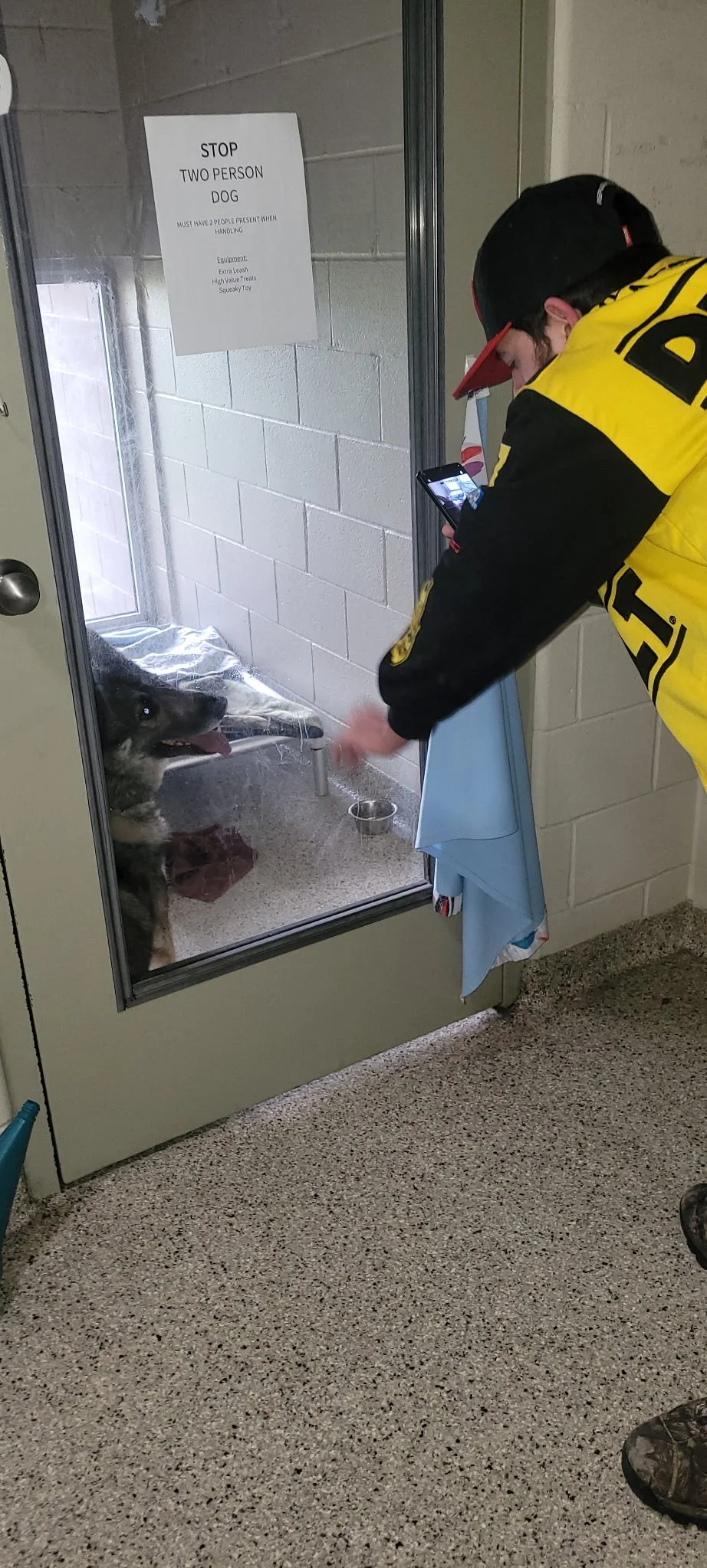 A man in a yellow and black jacket with a red and black cap is petting a dog inside a kennel through a glass door. The dog is sitting on a blanket on the floor of the kennel, with its tongue out. There is a sign on the door that reads: 'STOP TWO PERS