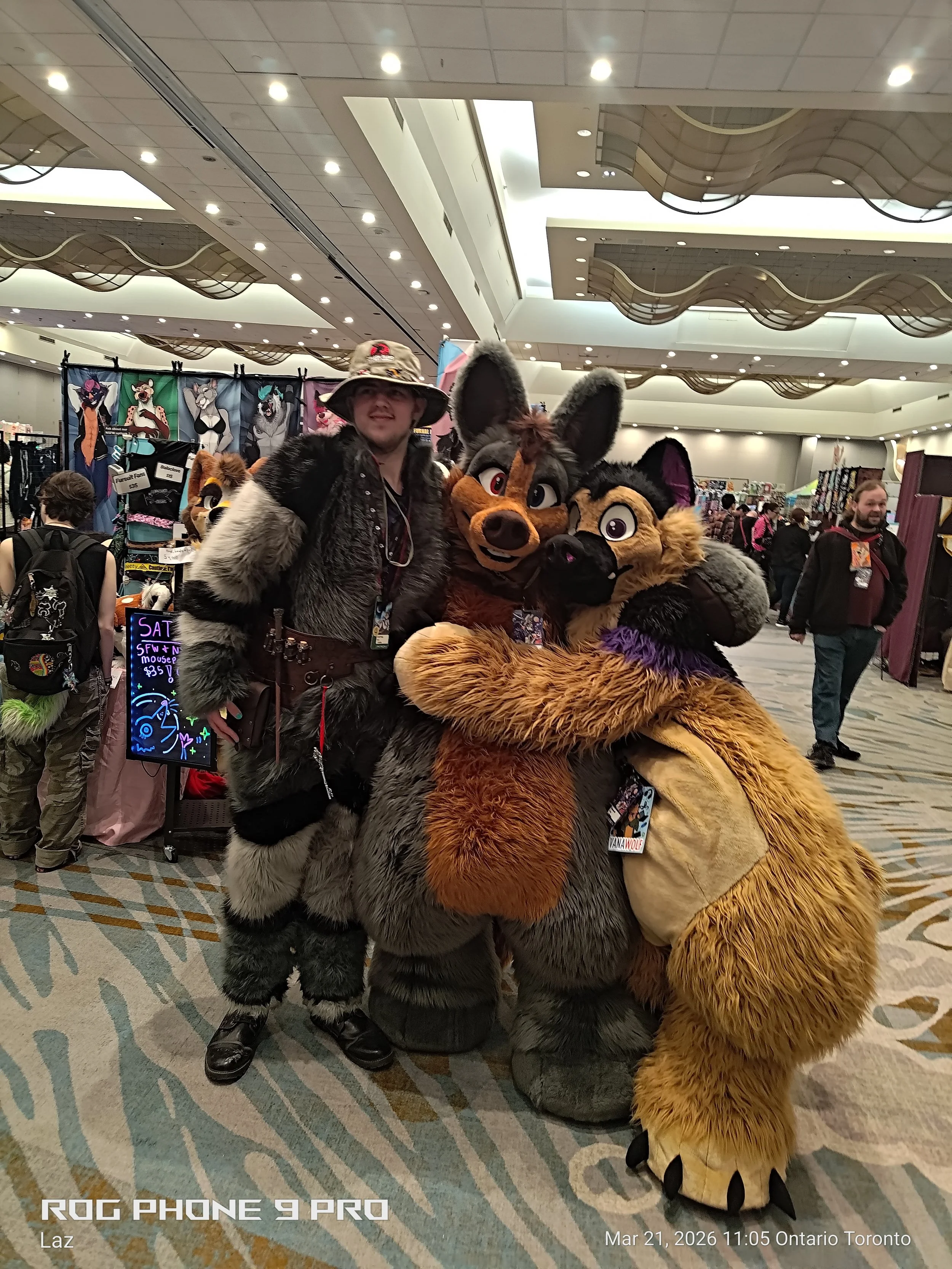 Three people dressed in furry costumes, including a wolf, a fox, and a raccoon, at a convention or event. They are posing together in a large indoor space with booths and merchandise in the background.