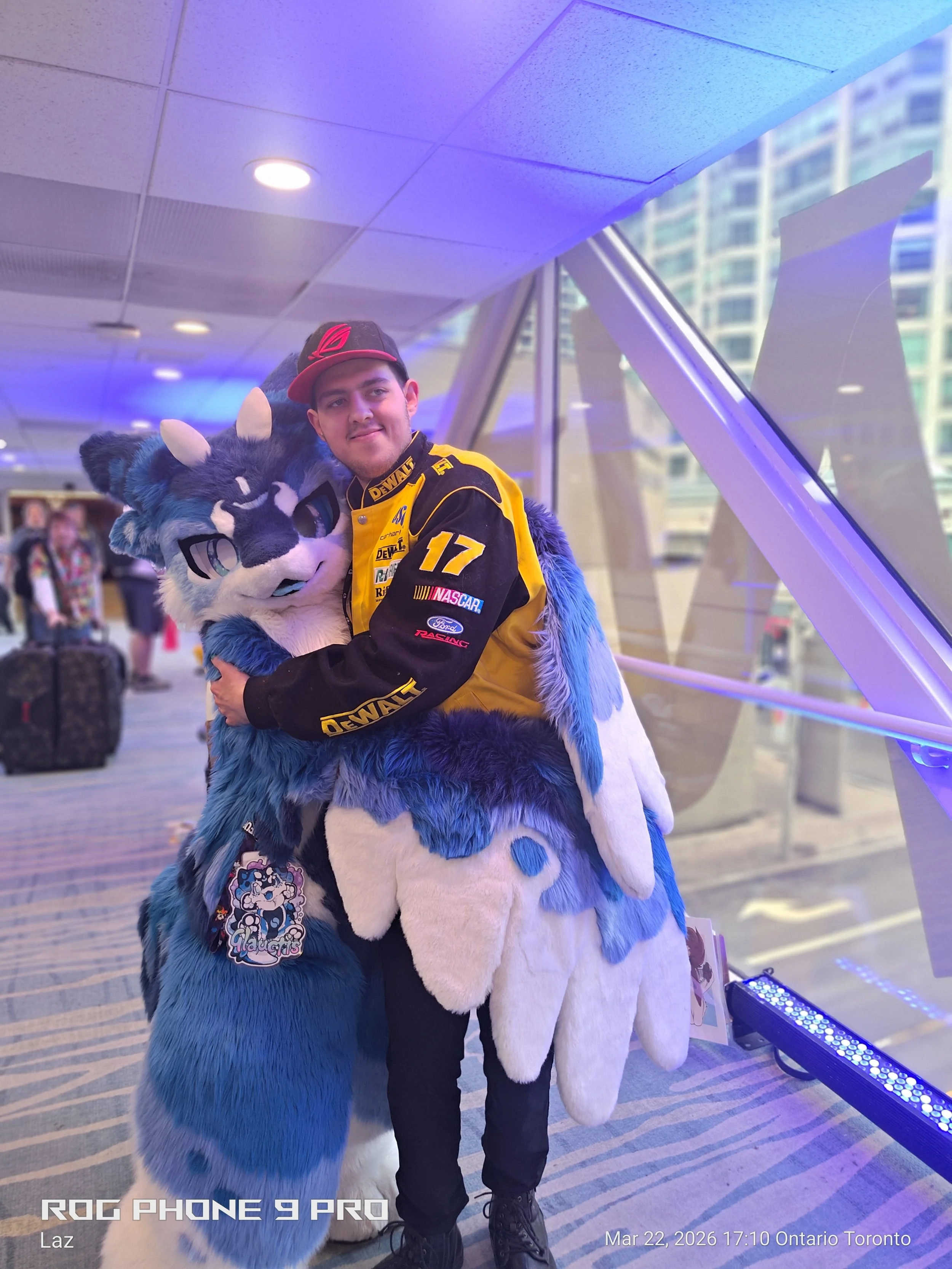 A person is hugging a person in a blue and white furry costume resembling a wolf or fox at an airport or convention center. The person in costume has a badge or sticker on their chest and is wearing a badge. The person in the costume is also wearing 