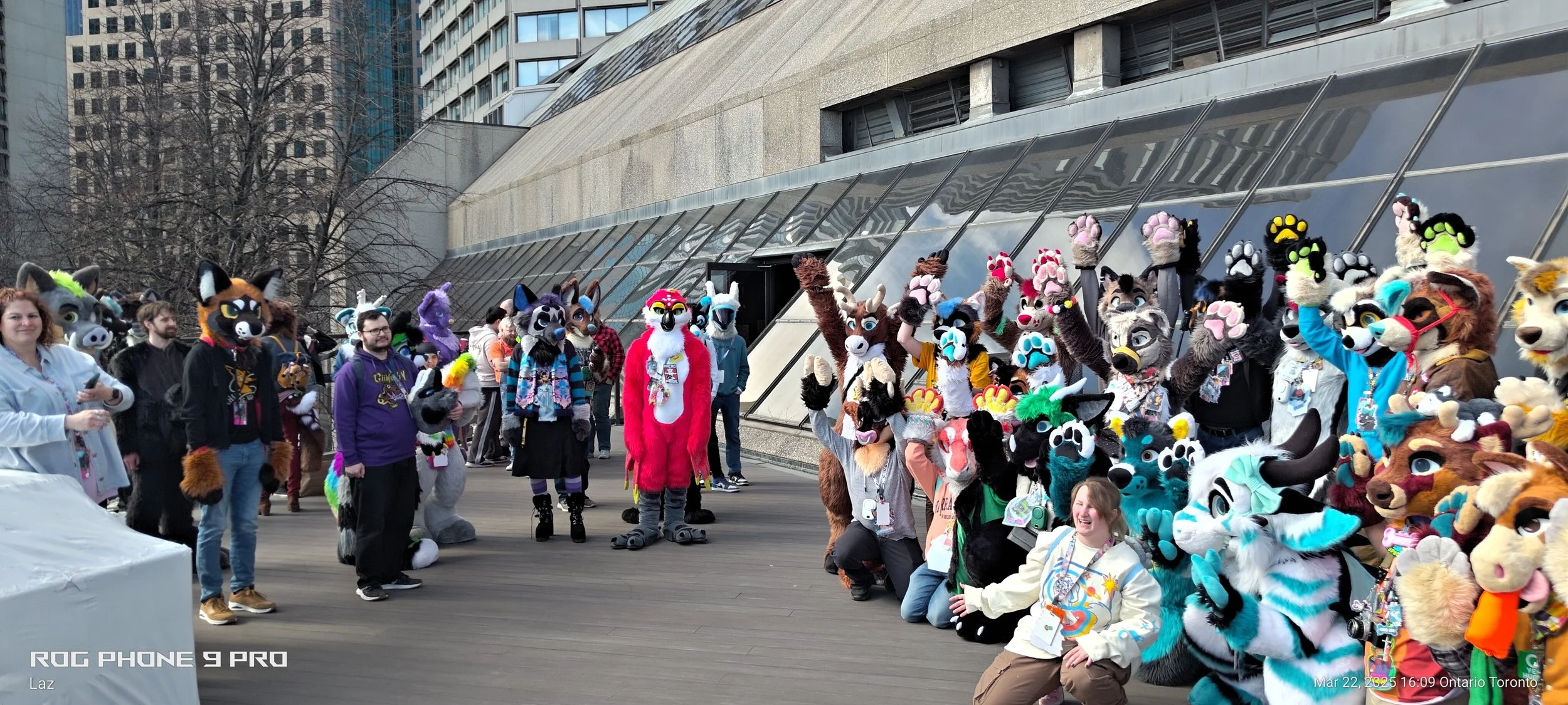 Group of people in colorful animal costumes, known as furries, gathered outdoors in front of tall buildings, some posing, smiling, and waving.