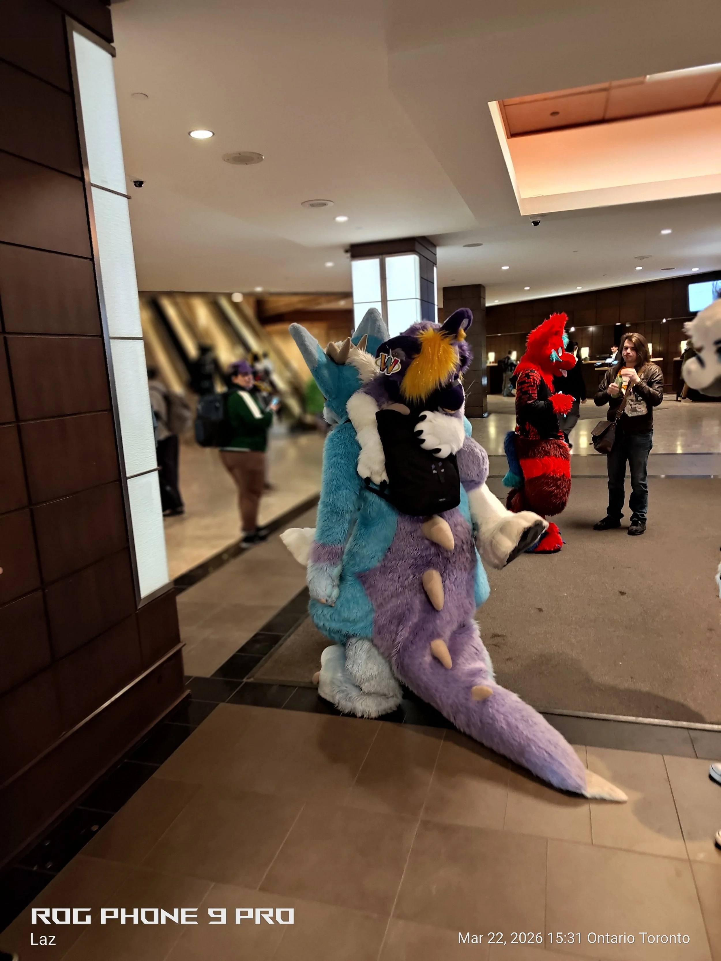 People dressed in colorful furry costume characters, one in blue and purple, and another in red and black, in what appears to be a hotel lobby. One person is wearing a gray jacket and holding a drink.