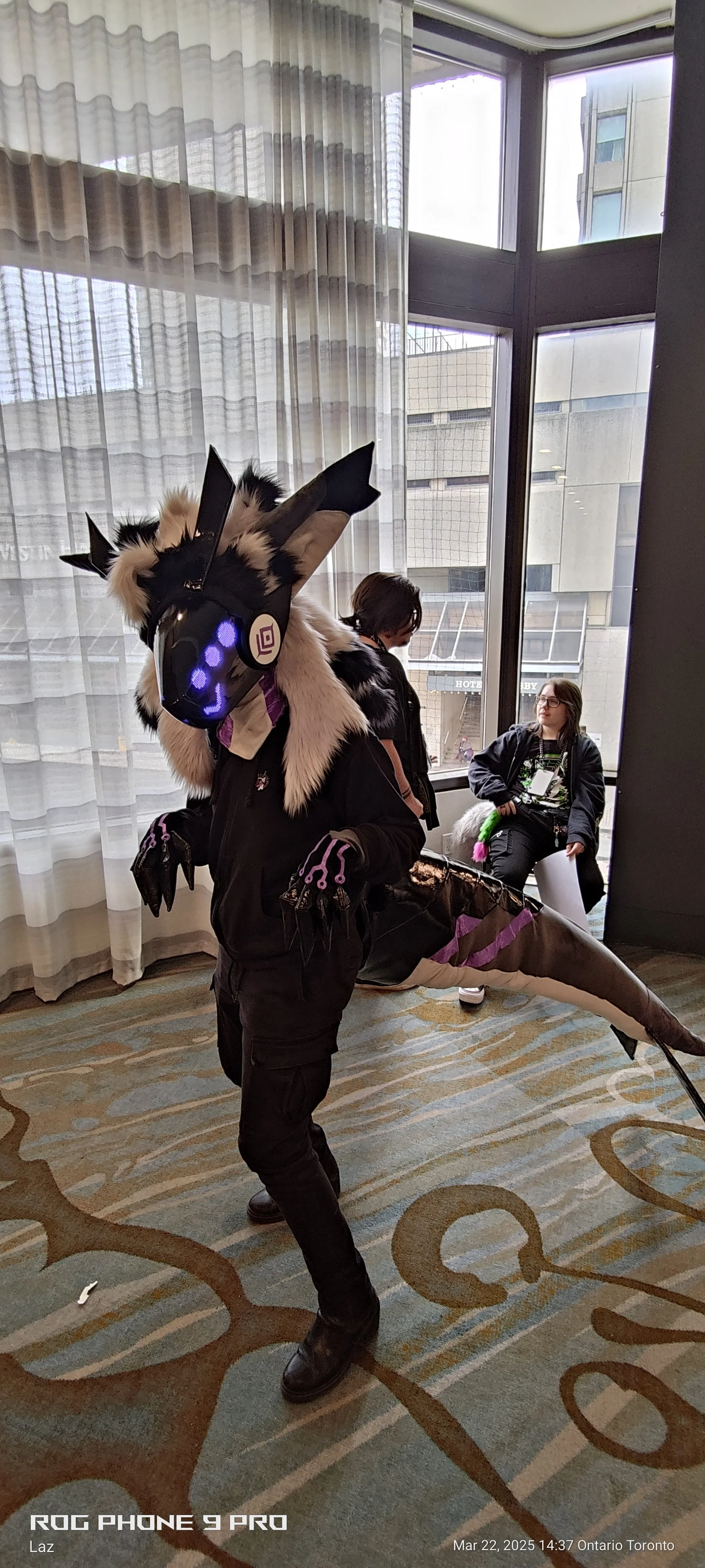 A person in a dragon costume with a glowing, robotic mask and fur accents, standing in a hotel lobby near large glass windows. Two other people are sitting and standing in the background, engaged in conversation.