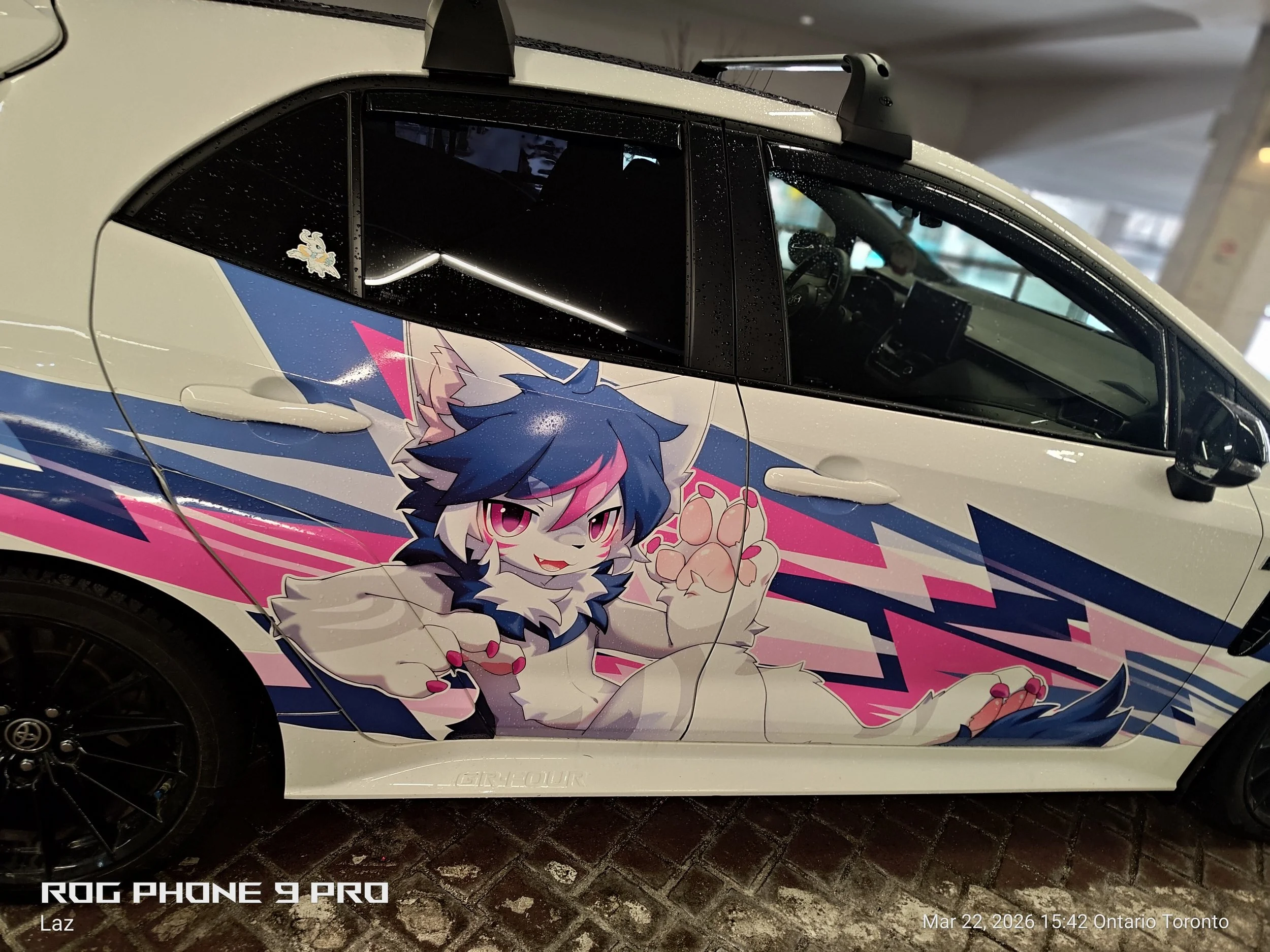 Close-up of a car with anime-style artwork featuring a cute wolf-like character with pink and blue hair, pink eyes, and paws, and a sticker of a white wolf on the rear window, parked indoors on a wet surface.