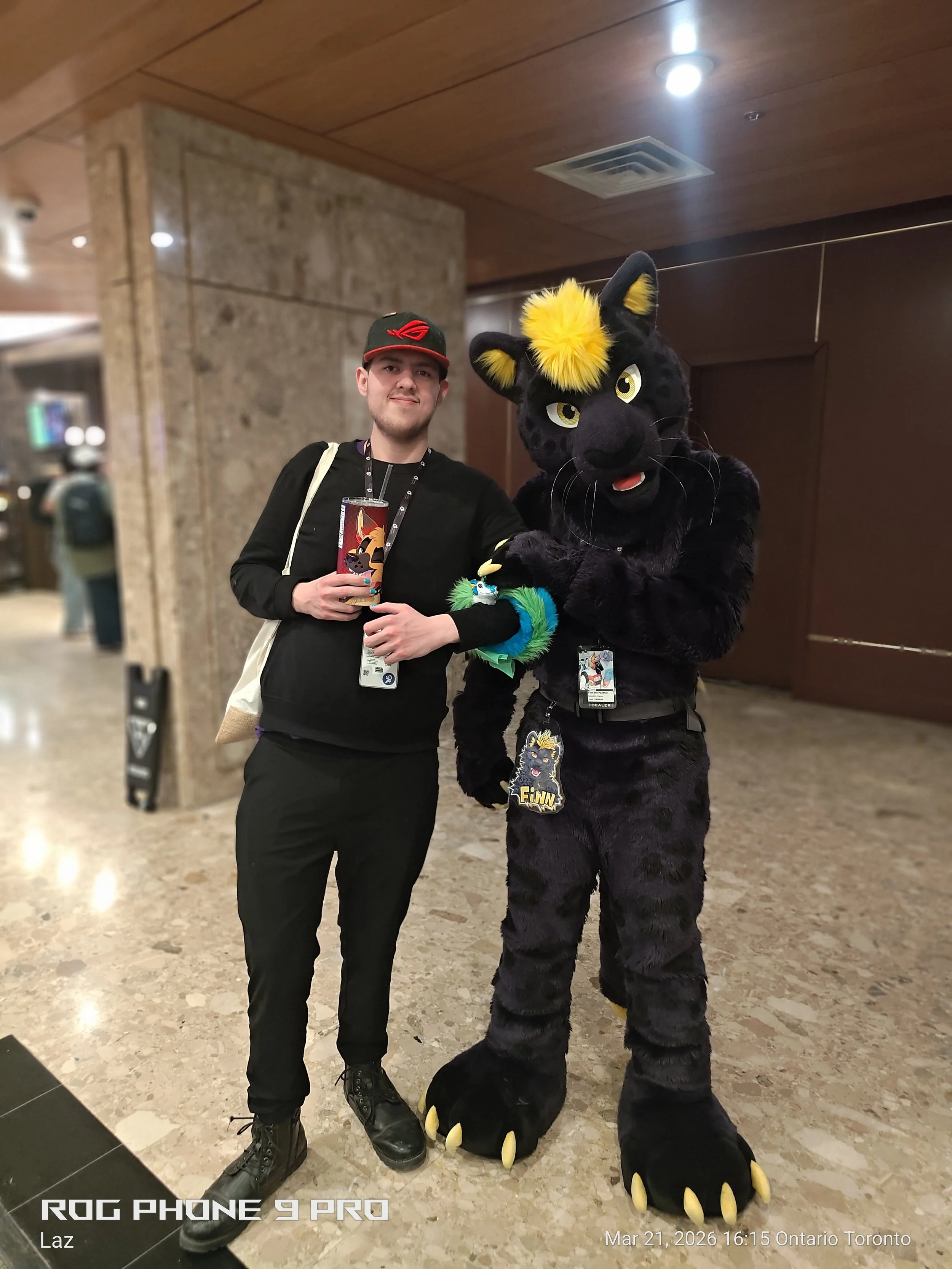 A man dressed in black holding a drink cup and a person in a black and yellow fuzzy panther costume posing together indoors