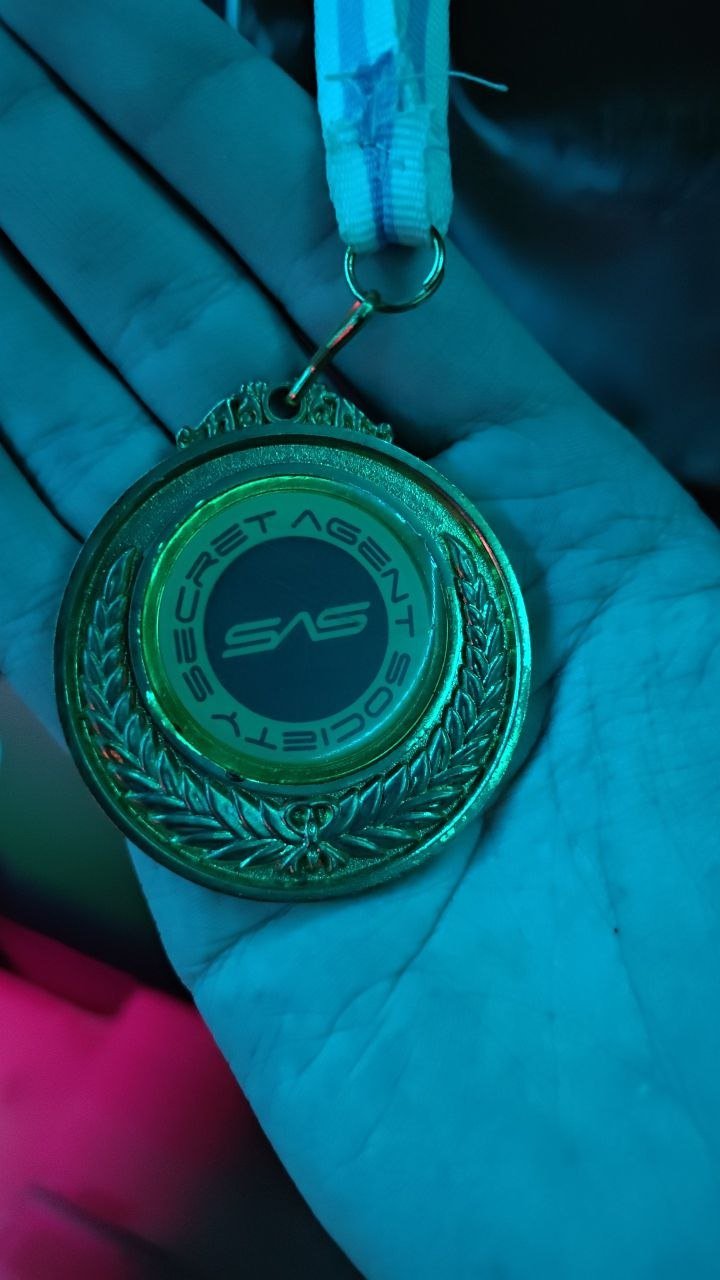 Close-up of a hand holding a keychain with a large circular badge that has the logos of the Secret Service and SAS. The badge is gold with laurel leaves design around the edge, and the background behind the badge is blue-green.