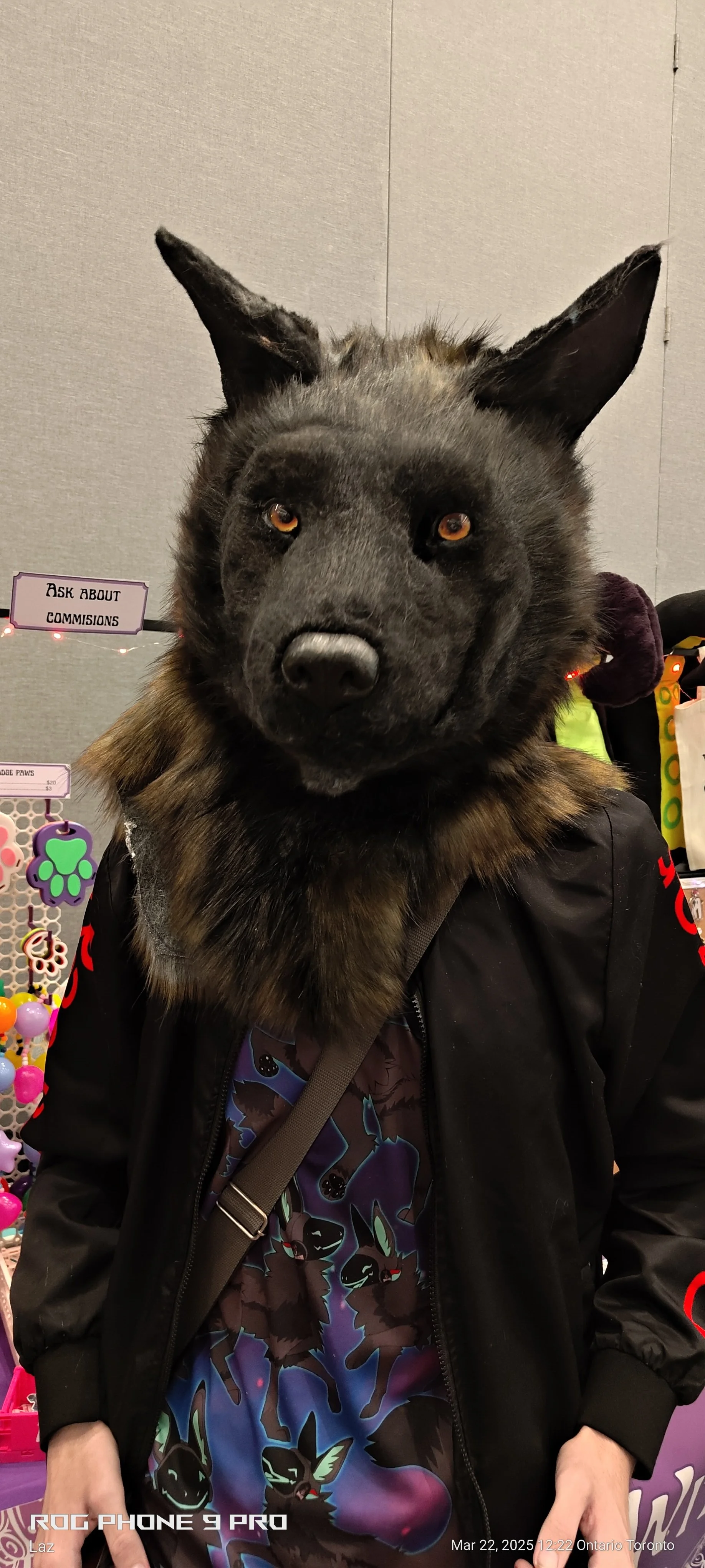 A person wearing a wolf mask with realistic furry fur and pointed ears, dressed in a black jacket over a Batman-themed shirt, standing in front of a display of colorful pet toys at a store, with a sign that reads 'Ask About Commissions' in the backgr