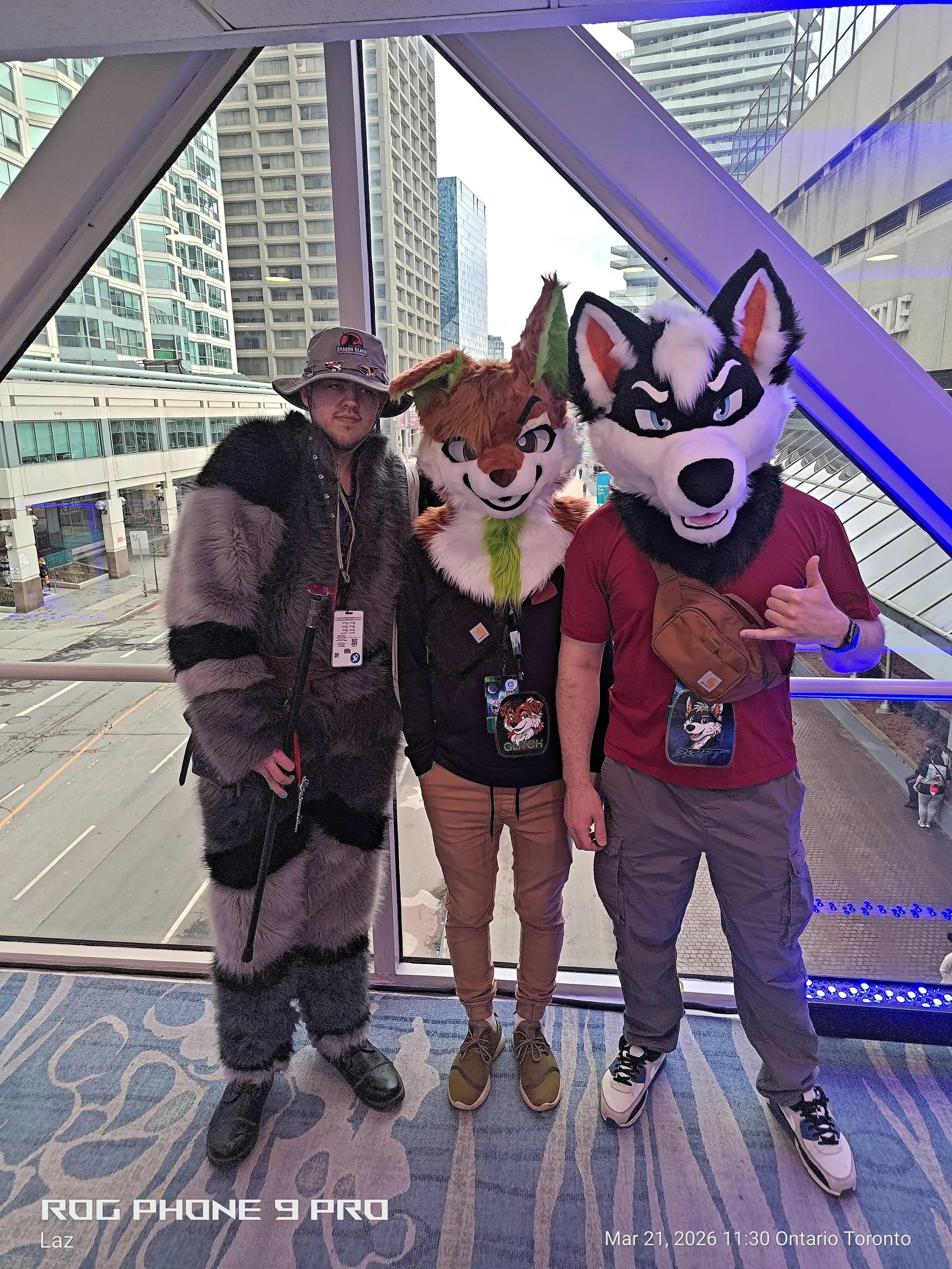 Three people standing together, two of them wearing animal costumes, in an urban setting with tall buildings visible through large windows behind them.