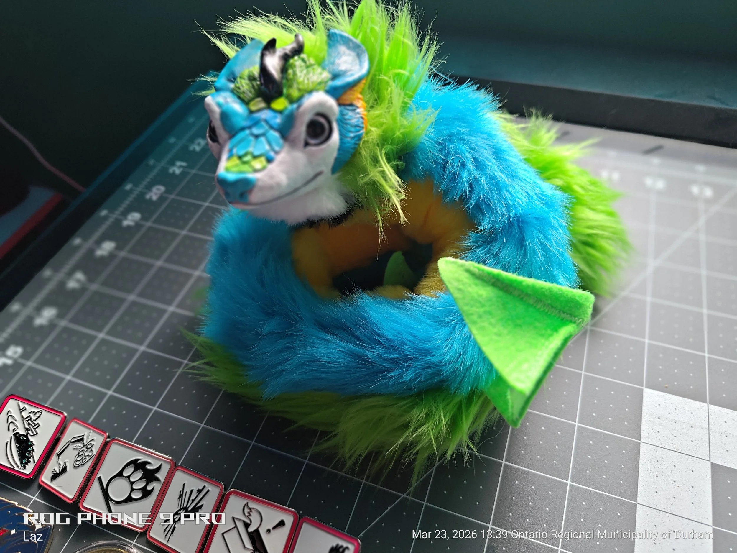 A plush toy resembling a colorful dragon with bright blue fur, green and yellow accents, and a detailed dragon face. It is placed on a cutting mat with playing cards visible in the lower left corner.