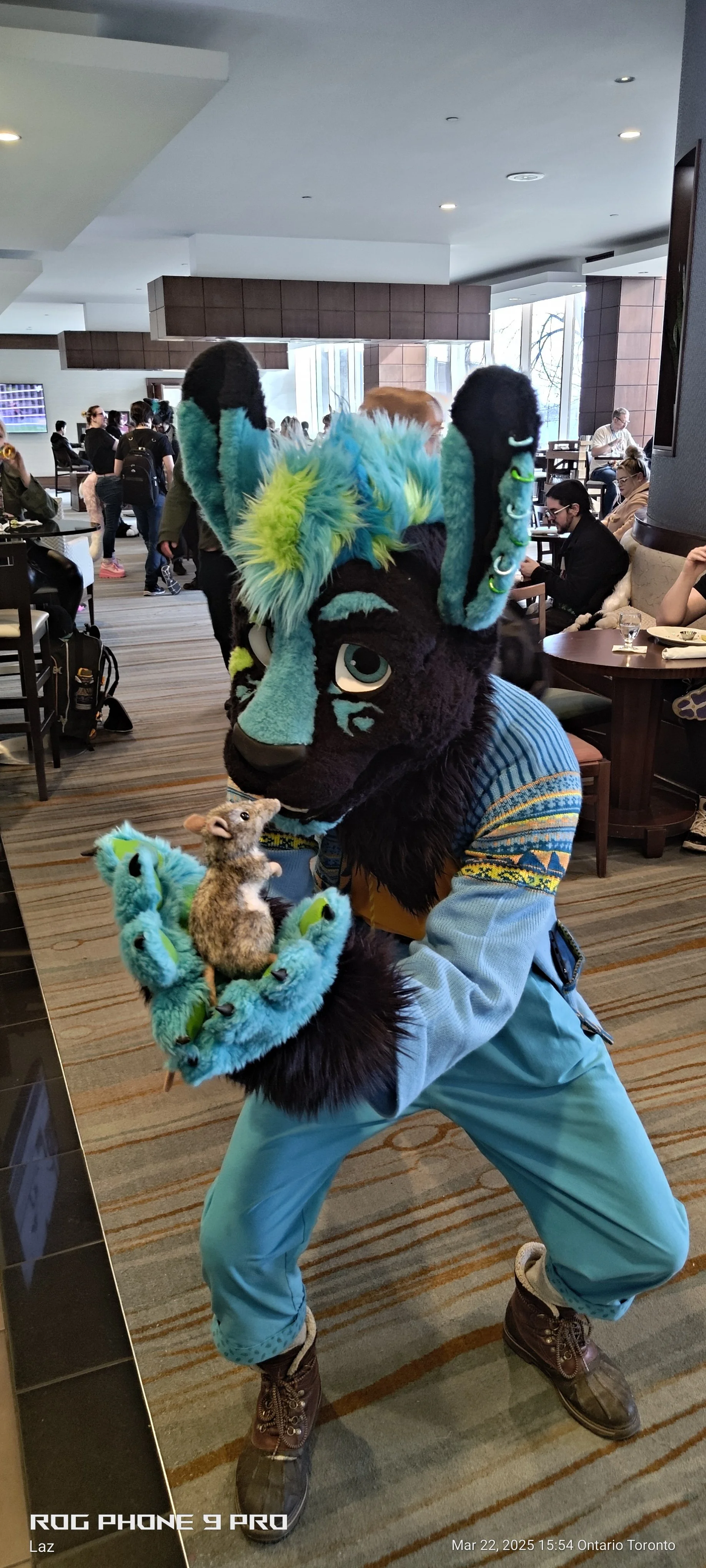 A person dressed in a colorful fursuit with a black face, teal, blue, and green features, holding a small stuffed animal resembling a rodent, inside a busy indoor setting.