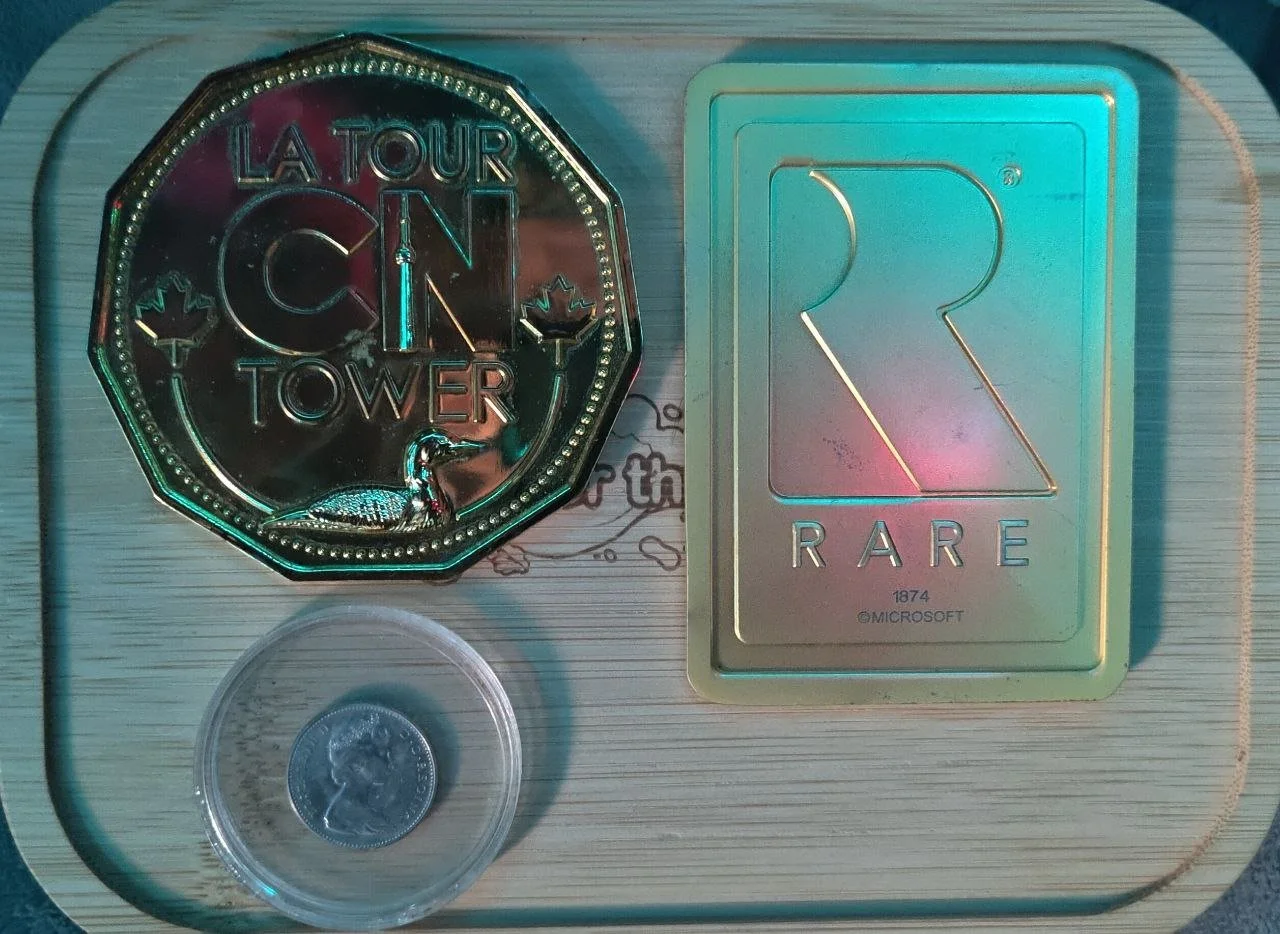 A collection of gaming-related items including a round coin with a portrait, a silver rectangular card with the letter R and the word RARE, a large black and silver badge with the words LA TOUR CN TOWER, and two small maple leaf icons.