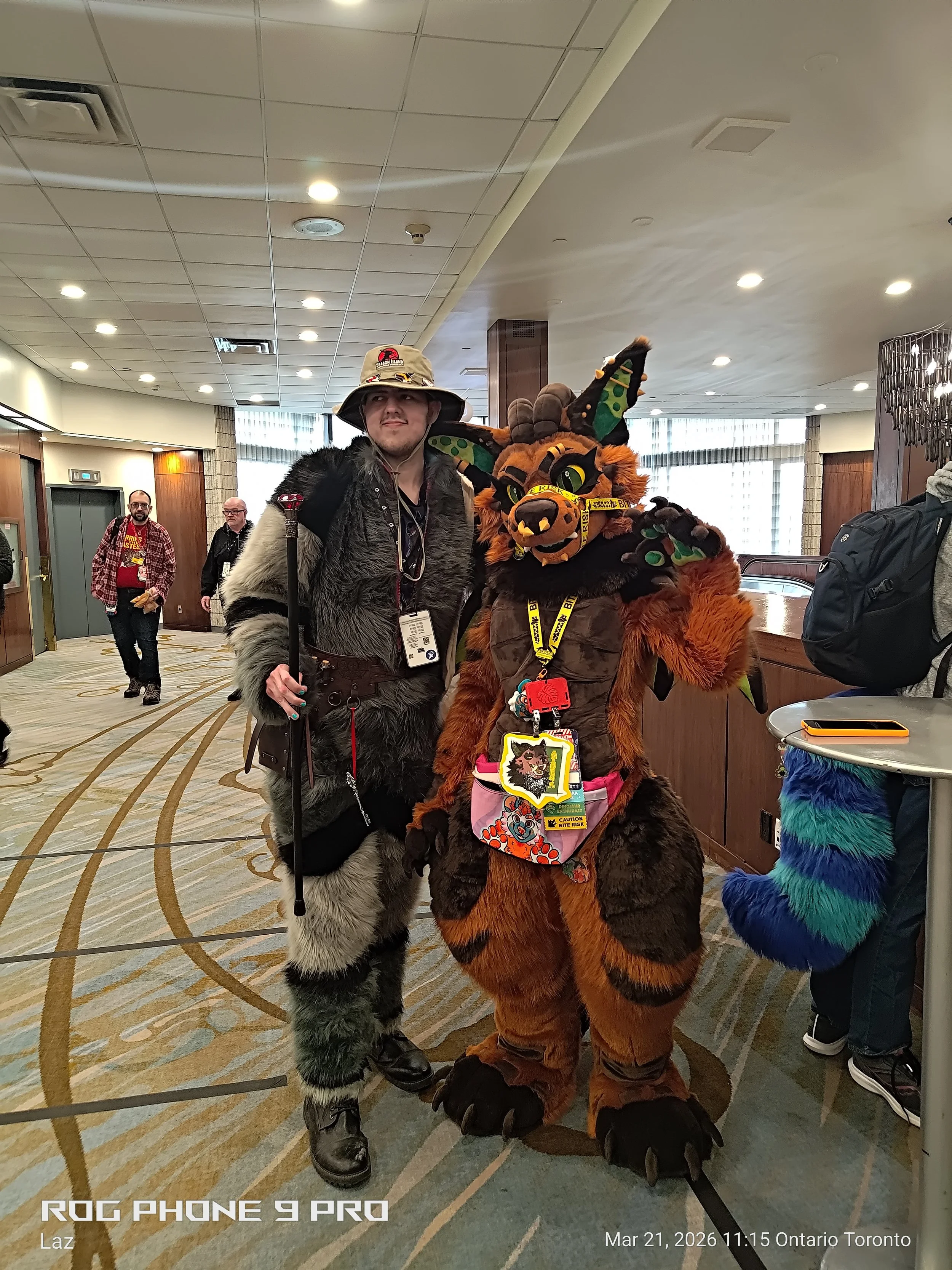 Two individuals in costume at a convention, one dressed as a furry wolf and the other as a furry tiger, standing in a hotel lobby with other attendees in the background.