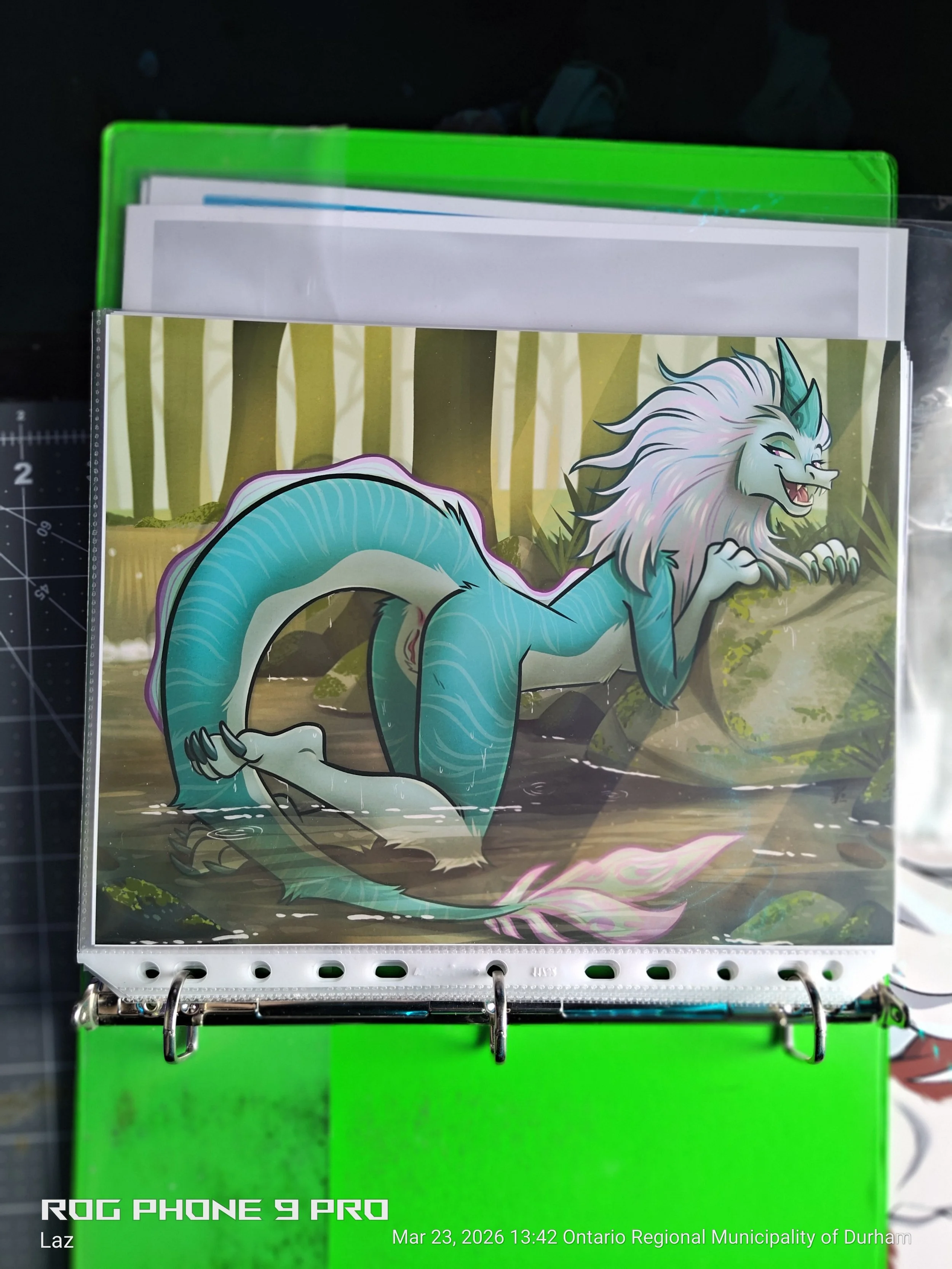 A plastic folder with a colorful illustration of a fantasy creature resembling a lion with a blue tail and a horn, set against a forest background, attached to a black surface with a ruler on the side and a timestamp.