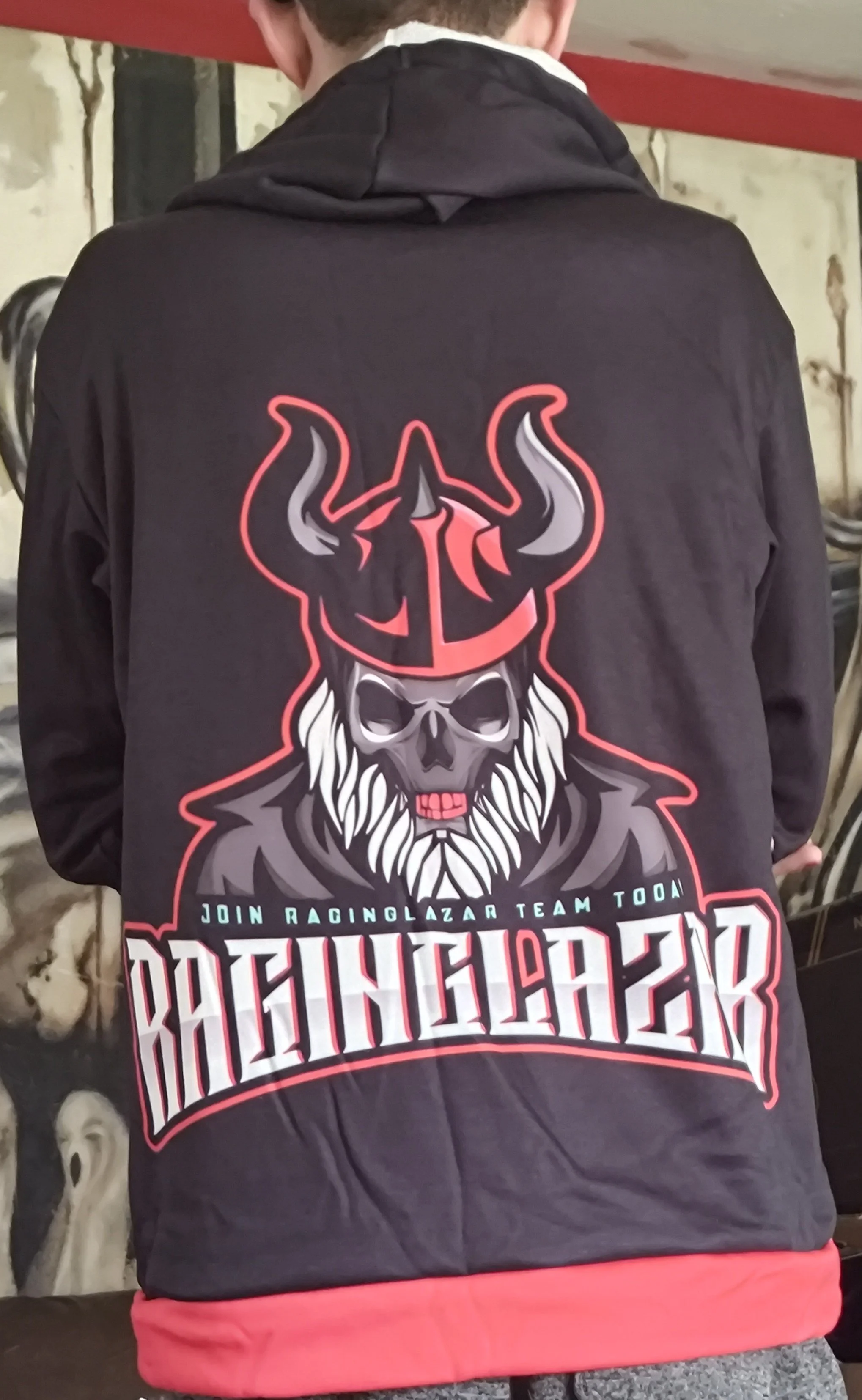 Back of a person wearing a black hoodie with a graphic design of a skull with a beard and horns, with text that reads 'Join RacingLazar Team Today' and 'FightingBack' in red and white lettering, standing indoors in front of a wall.