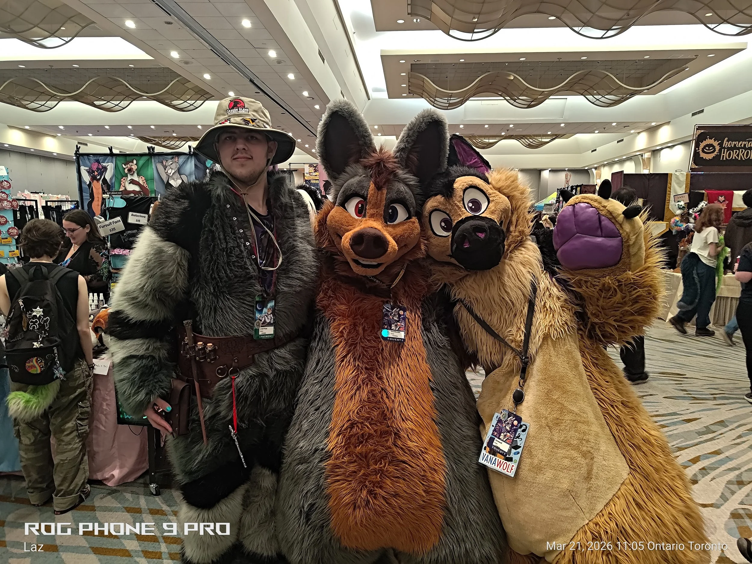 People at a convention, including two individuals in large animal costumes resembling wolves, with one person in plain clothes wearing a hat and furry jacket, inside a spacious hall with booths and decorations.