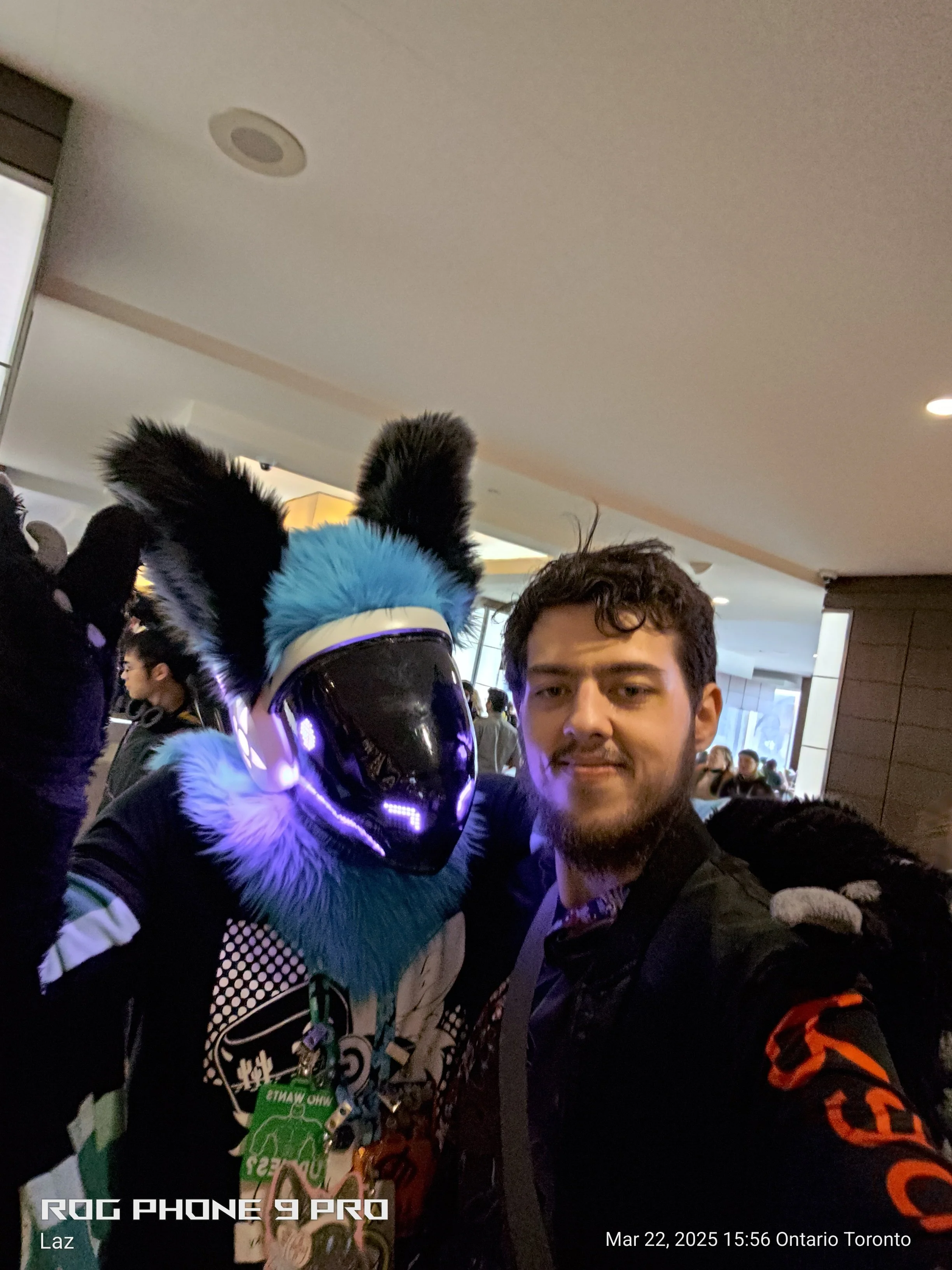 A person wearing a futuristic helmet with flashing lights and furry ears, standing next to a man in a crowded indoor setting.