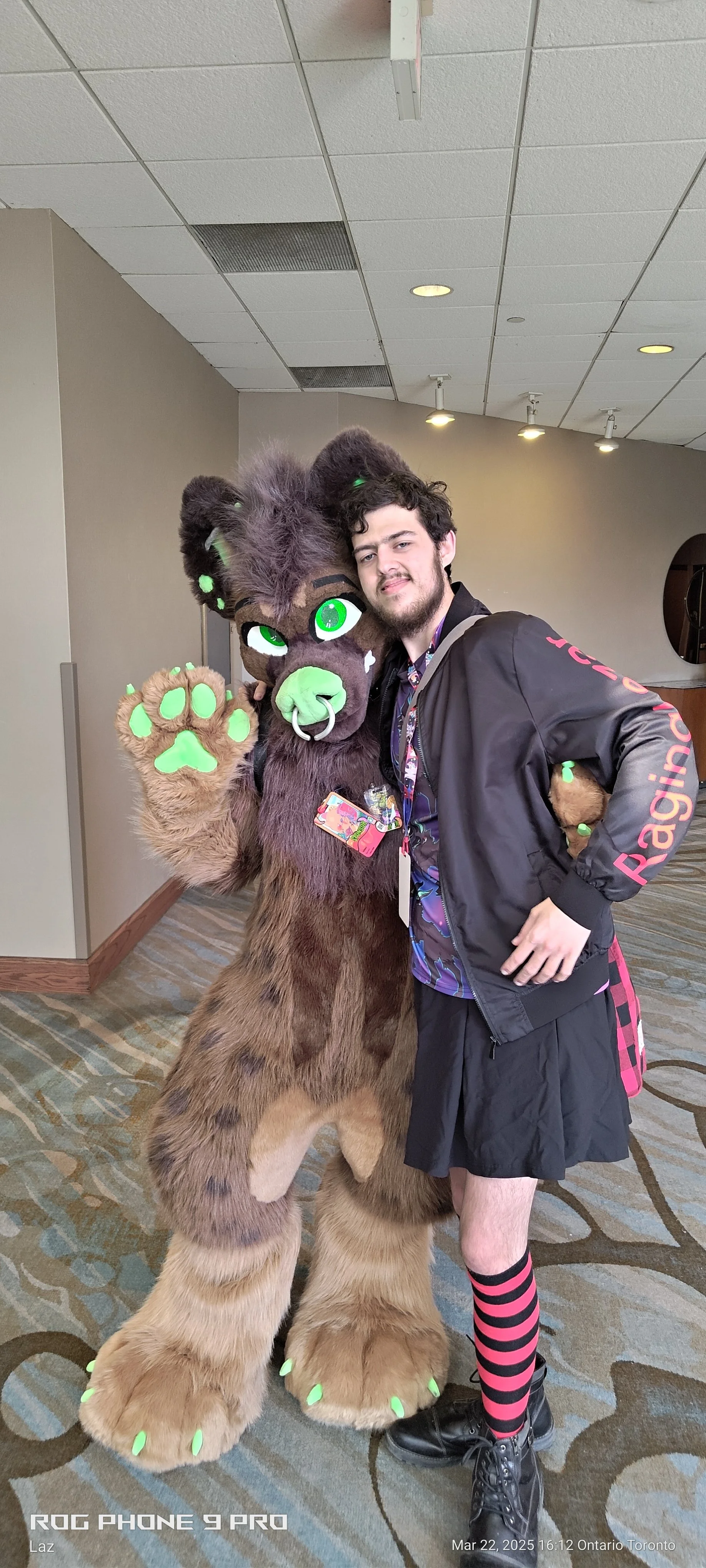 A person in a black and pink striped sock and black shoes standing next to a person in a furry costume with large green eyes and green paw pads, posing together indoors.