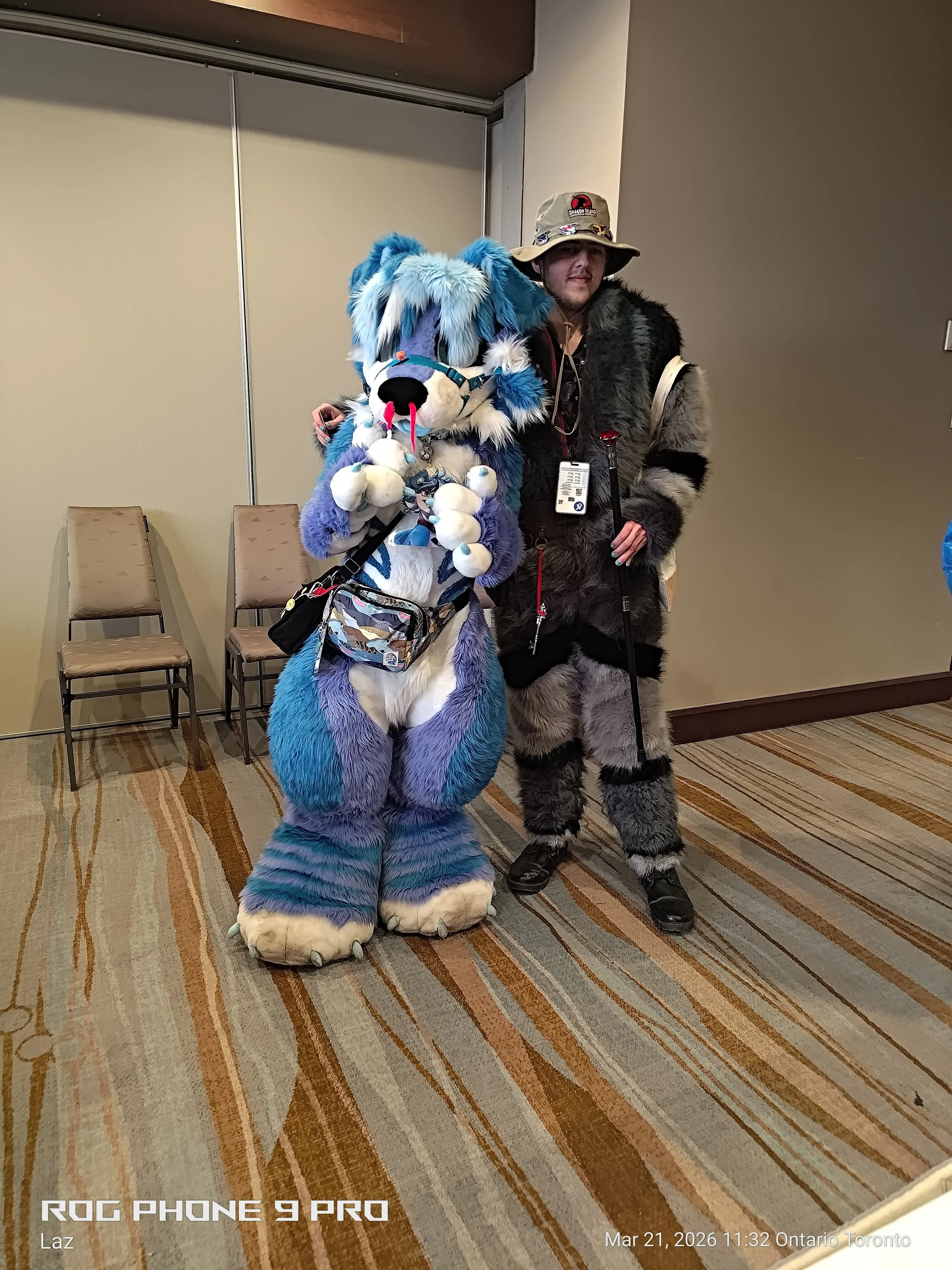 Two people in costume standing against a wall, one dressed as a furry blue and purple creature with a large plush toy, and the other dressed in a bear costume with a hat, holding a cane.