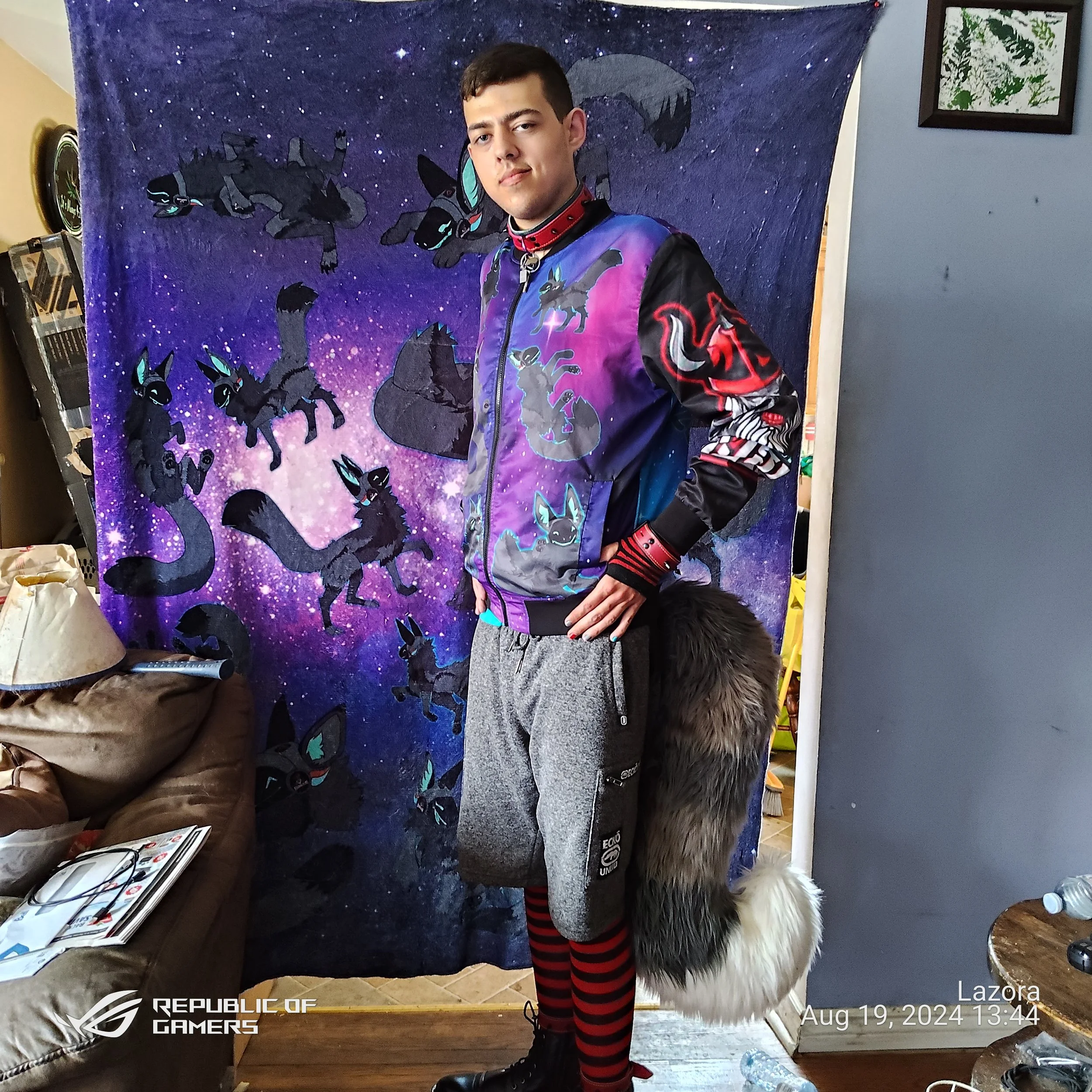 Young man standing indoors in front of a dark blue banner with a galaxy or space design and cartoon characters resembling black cats with teal eyes and red accents. He wears a colorful jacket with space and anime-themed graphics, red and black stripe
