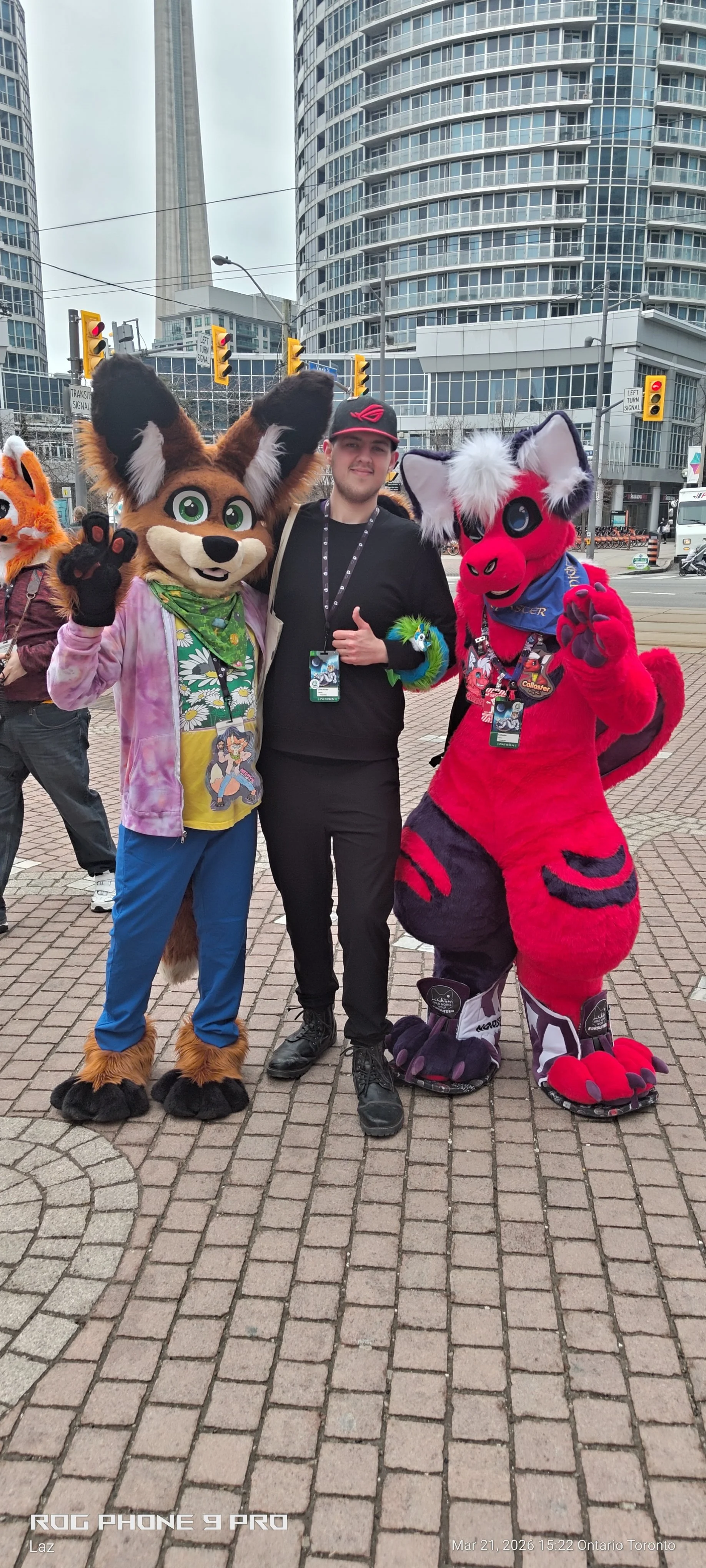 A man standing between two people in colorful animal costumes, in an urban setting with tall buildings, traffic lights, and a city street.