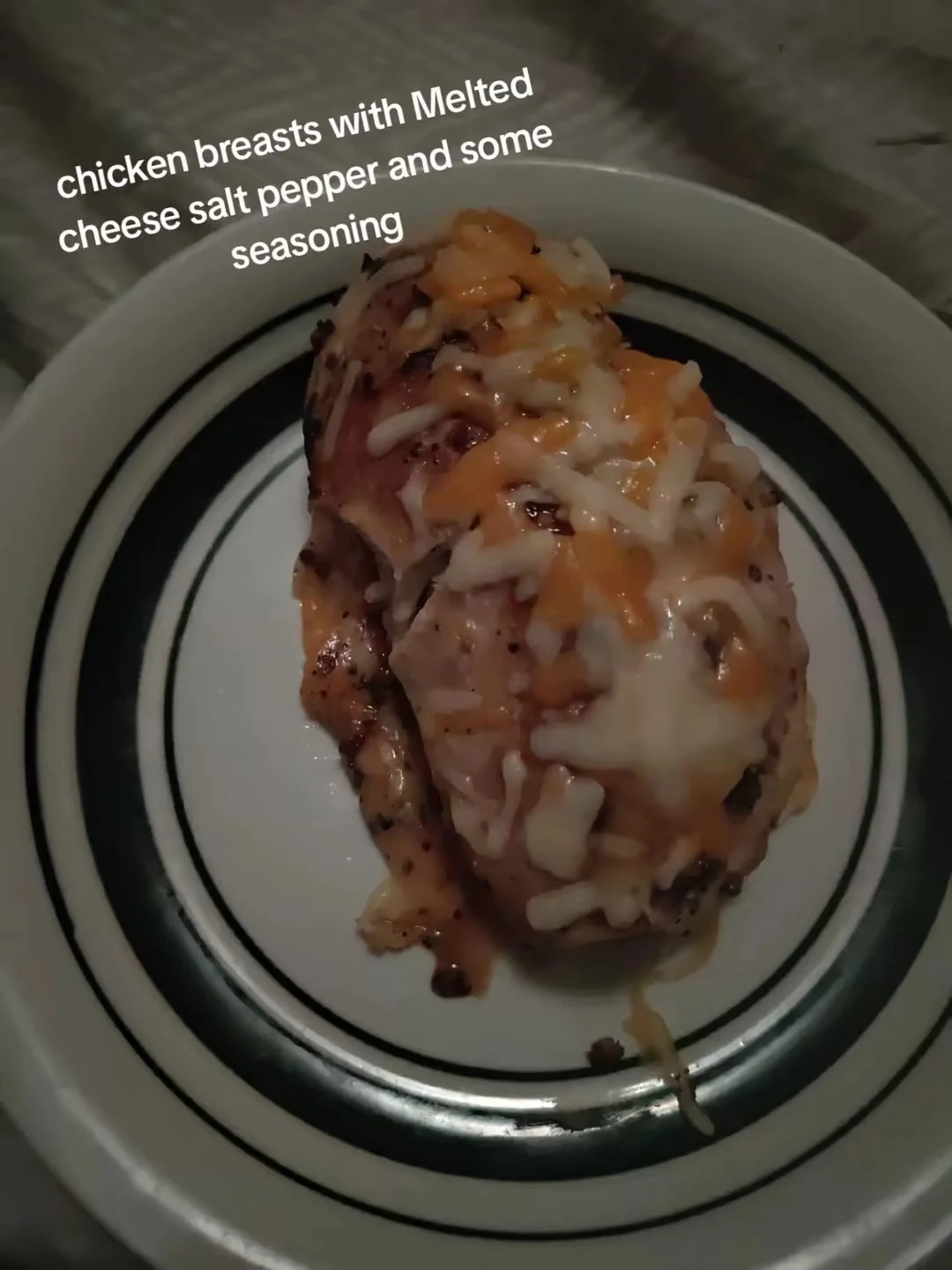 Two cooked chicken breasts topped with melted cheese, salt, pepper, and seasoning on a white plate with black decorative lines.