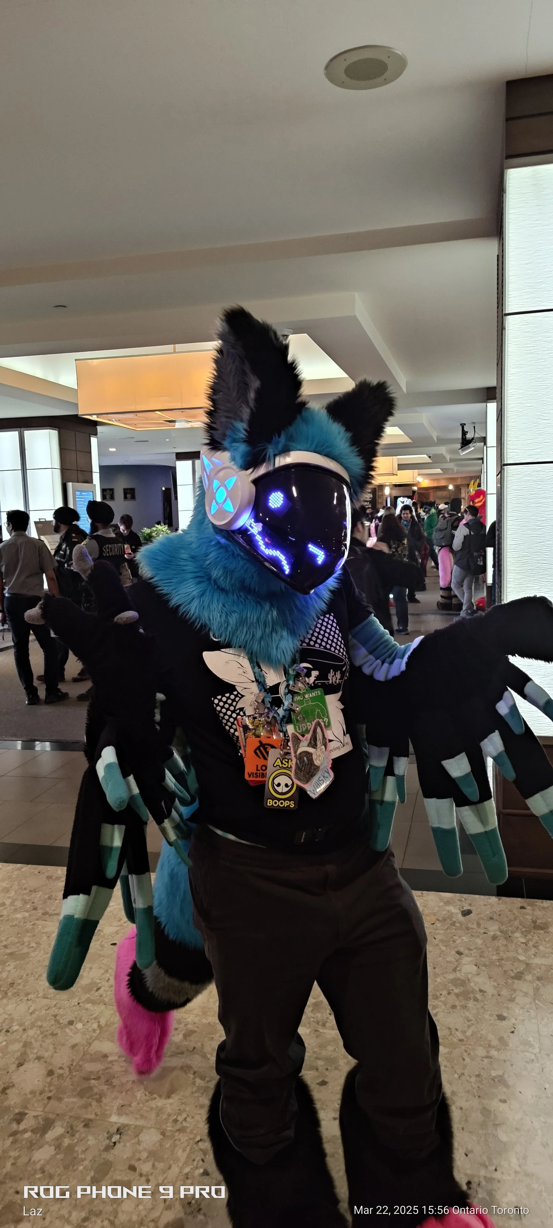 Person in a blue fluffy costume with a black helmet featuring glowing blue symbols, carrying multiple badges and necklaces, at an indoor event with other people in the background.