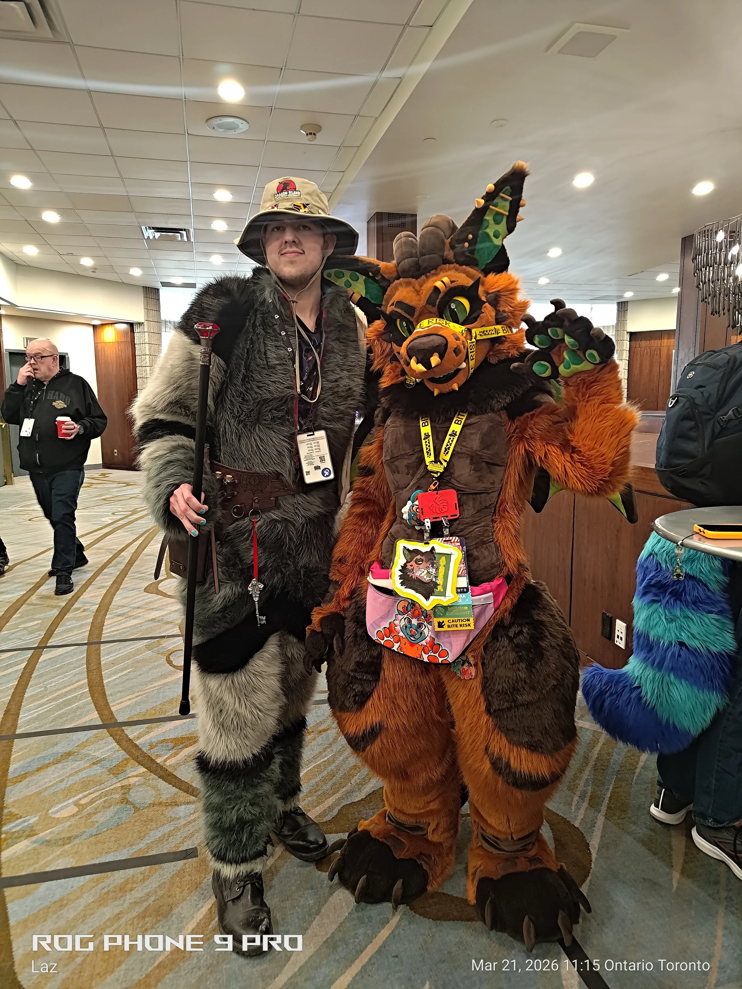 Two people in costume at a convention. One person is dressed as a furry wolf and the other as a dragon with horns and green face claws. They are posing together inside a building with a tiled ceiling and wooden wall panels.