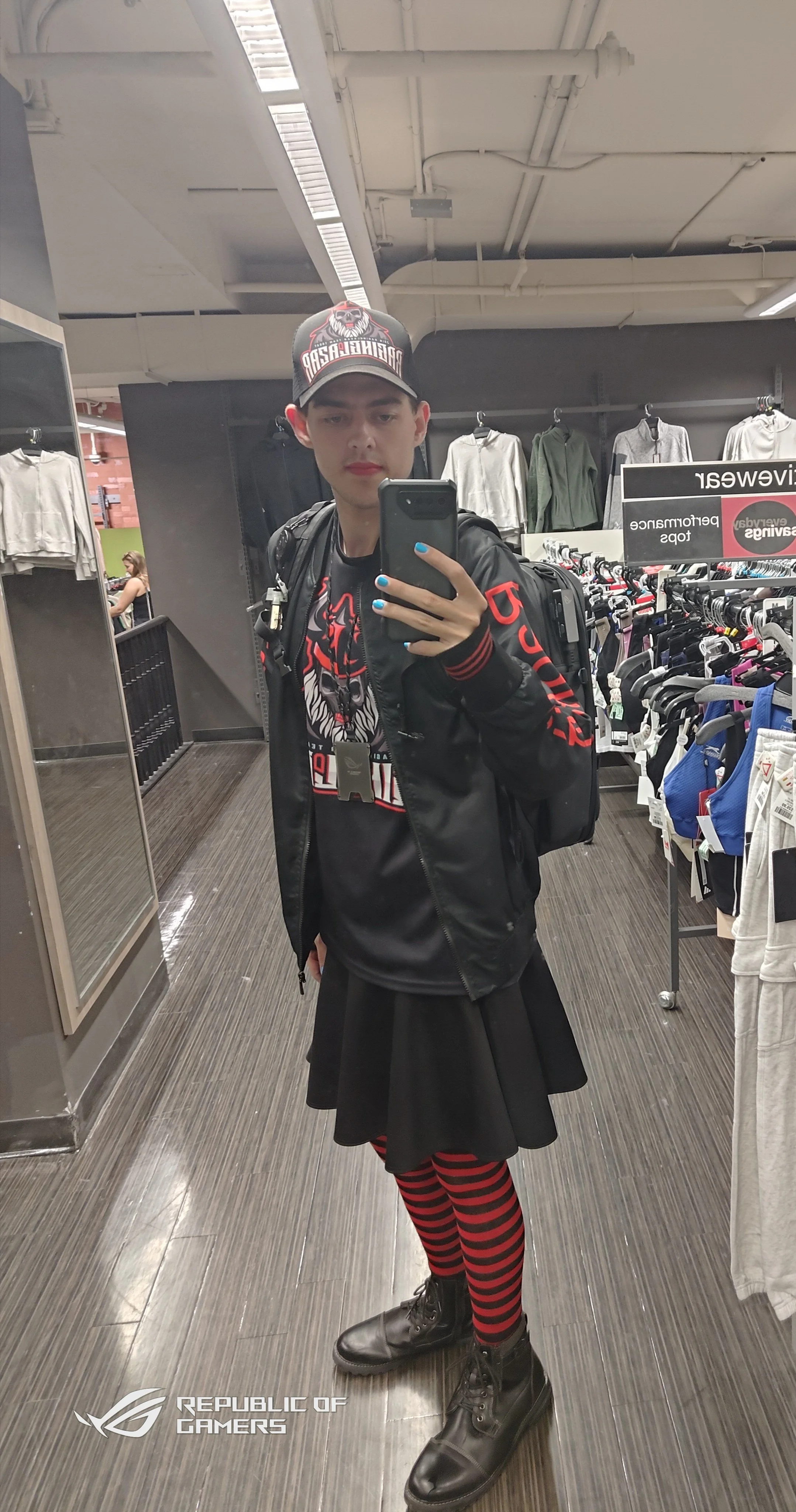 A young man taking a selfie in a clothing store wearing a black jacket, a graphic t-shirt, a black and red cap, black boots, a black backpack, and red and black striped tights, with clothing racks in the background and a logo for 'REPUBLIC OF GAMERS'