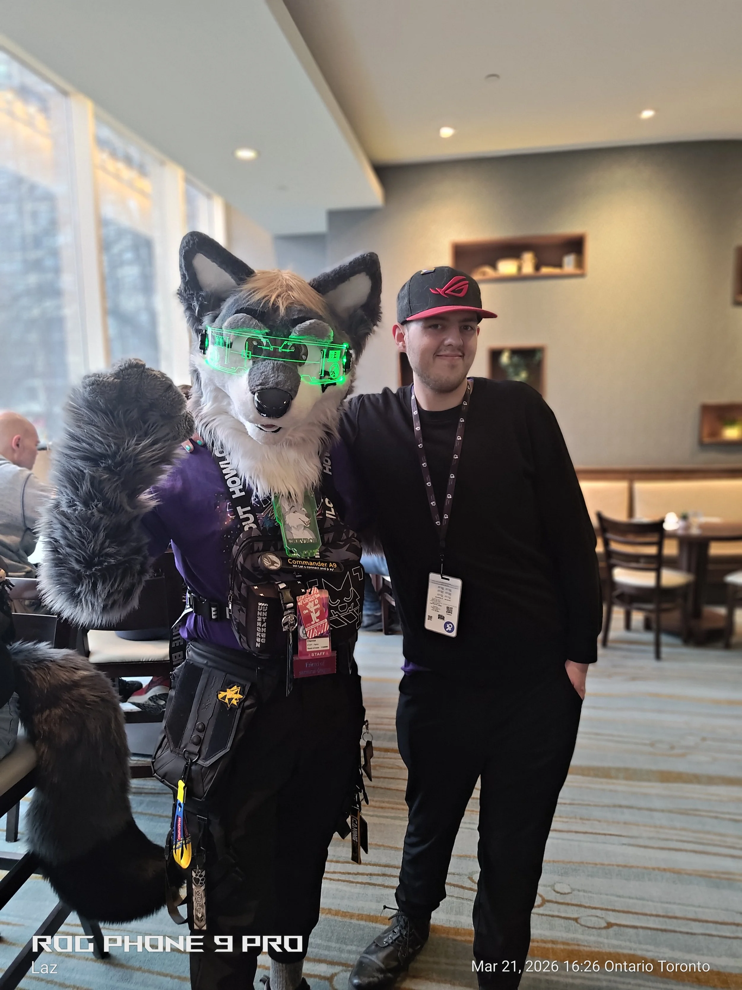 A person in a wolf costume with glowing green glasses standing beside a man in a black shirt and cap, both posing together indoors, with shelves and tables in the background.
