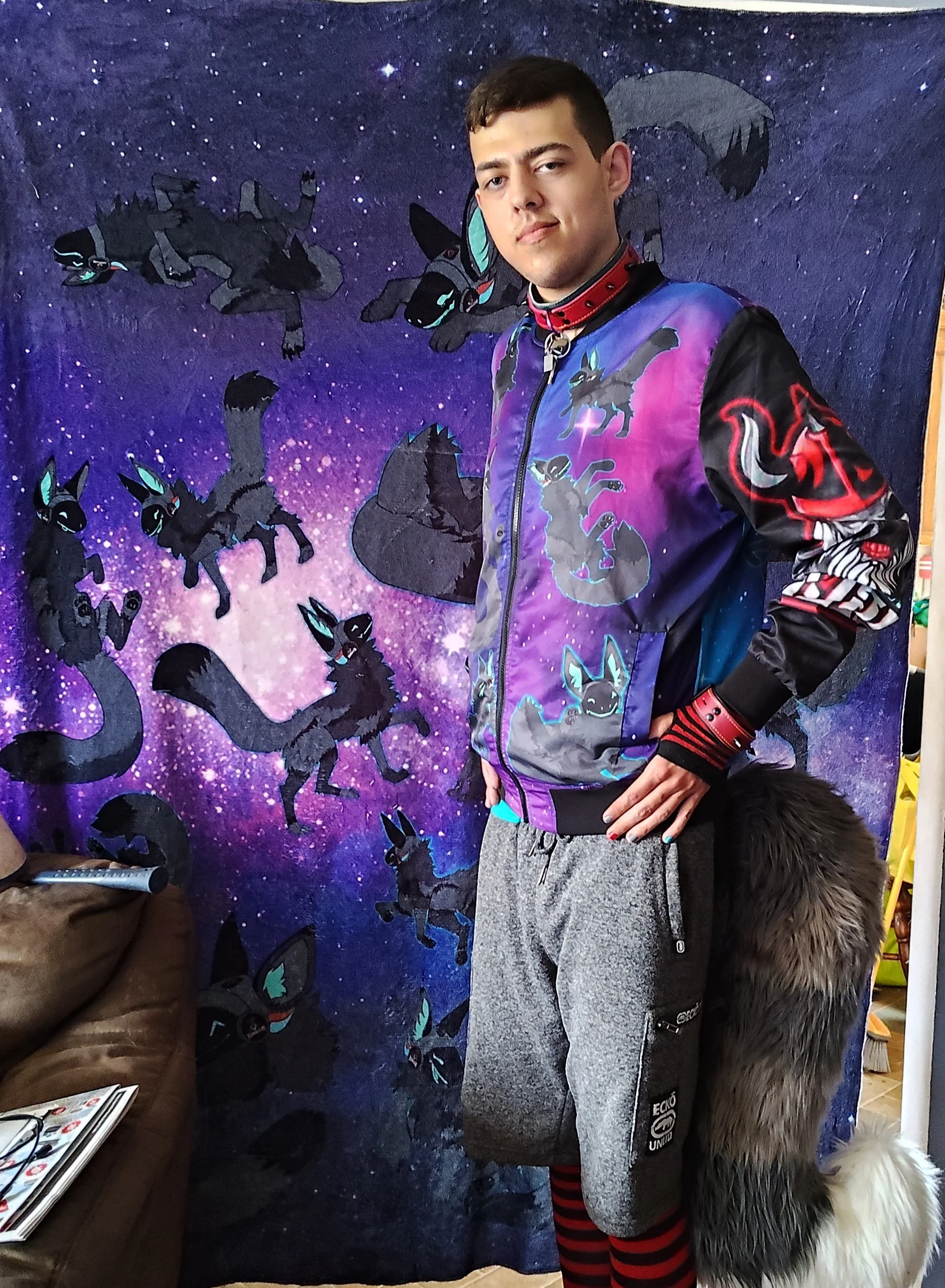 A young man with short dark hair stands in front of a painted backdrop featuring black lightning cats with blue eyes, a starry purple galaxy background, and silhouettes of flying black cats with blue eyes. The young man wears a colorful jacket with a