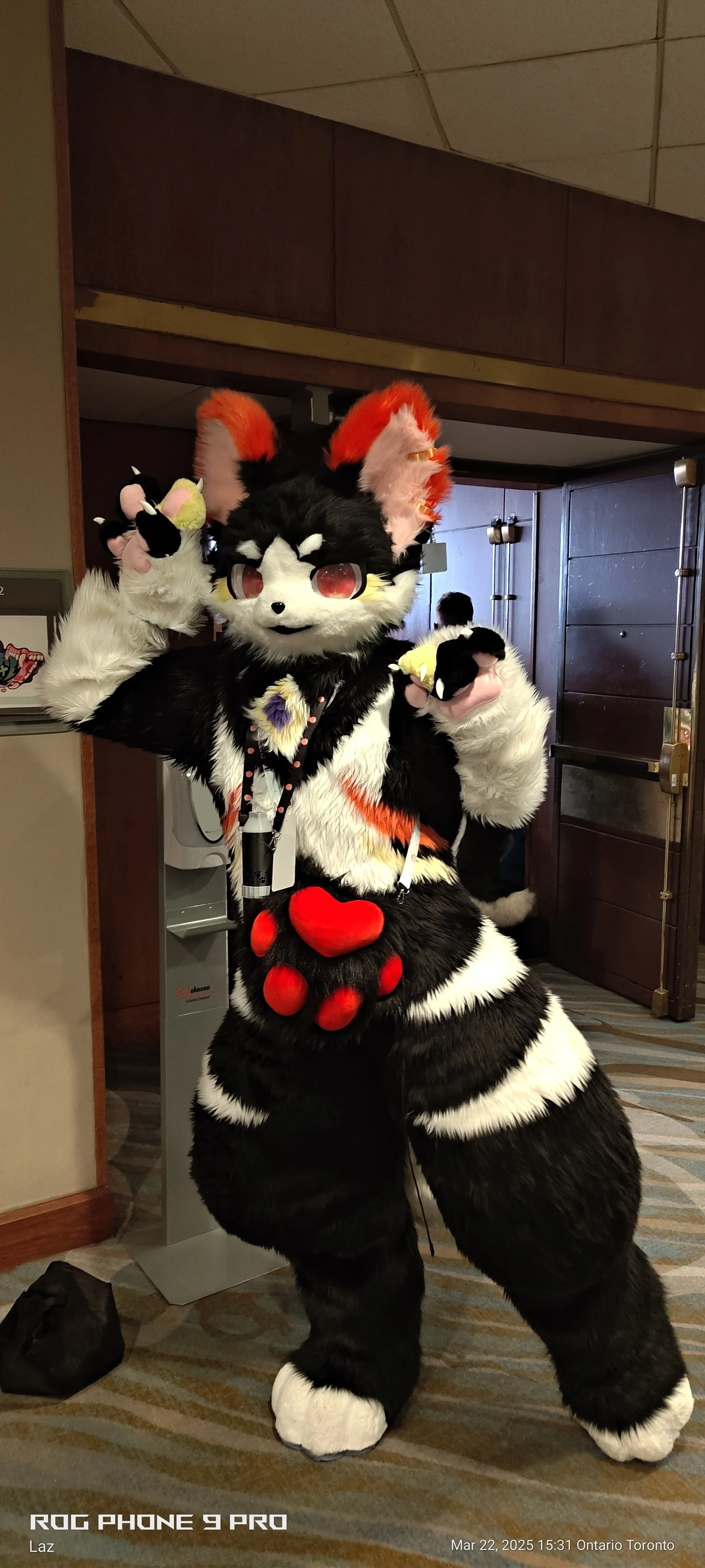 A person in a detailed wolf or fox mascot costume with large red and white ears, big red eyes, and red paw pads on the paws, standing indoors at a convention or event.