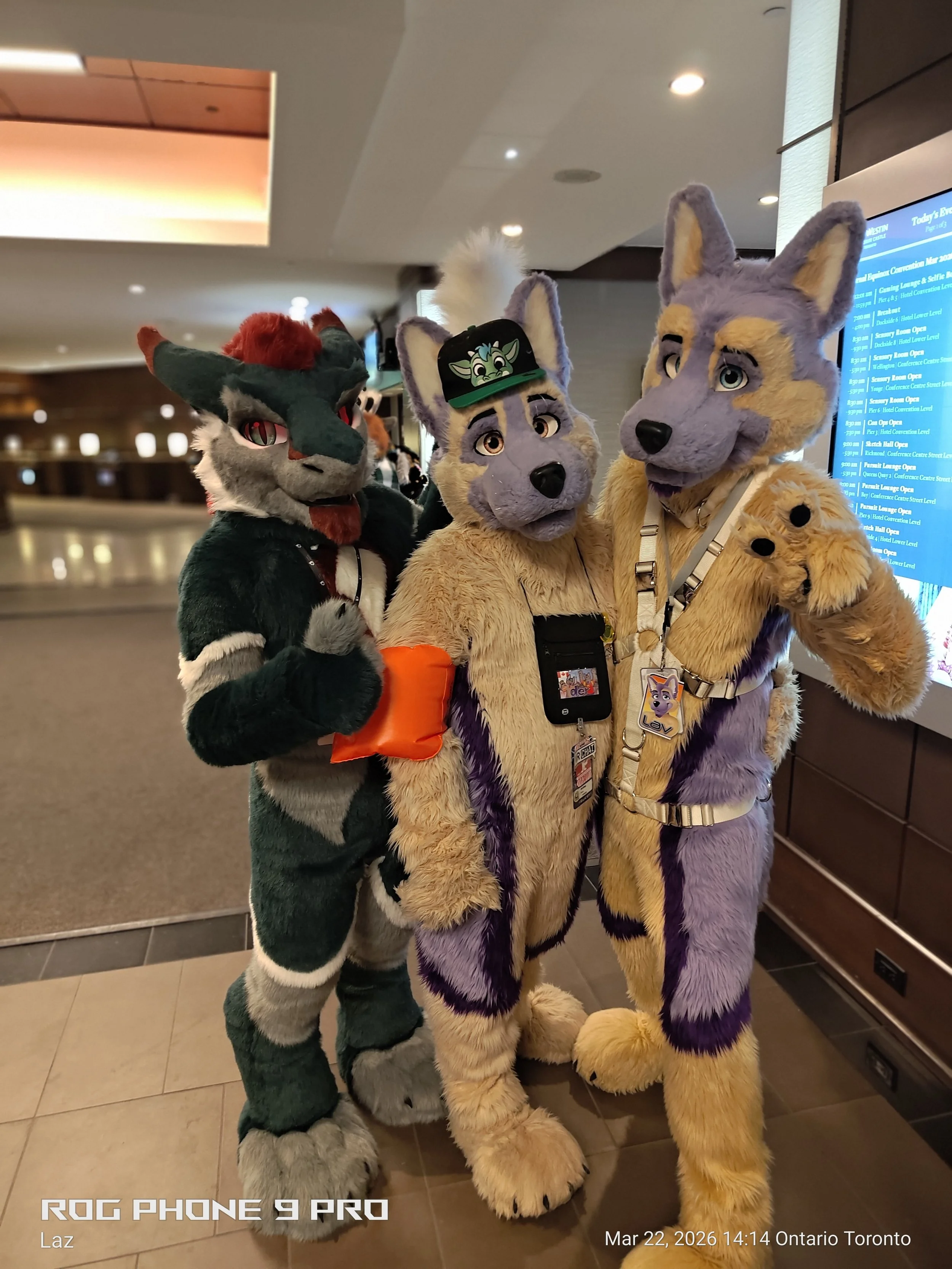 Three people dressed in colorful furry animal costumes posing together indoors, with a digital display screen in the background showing event information.