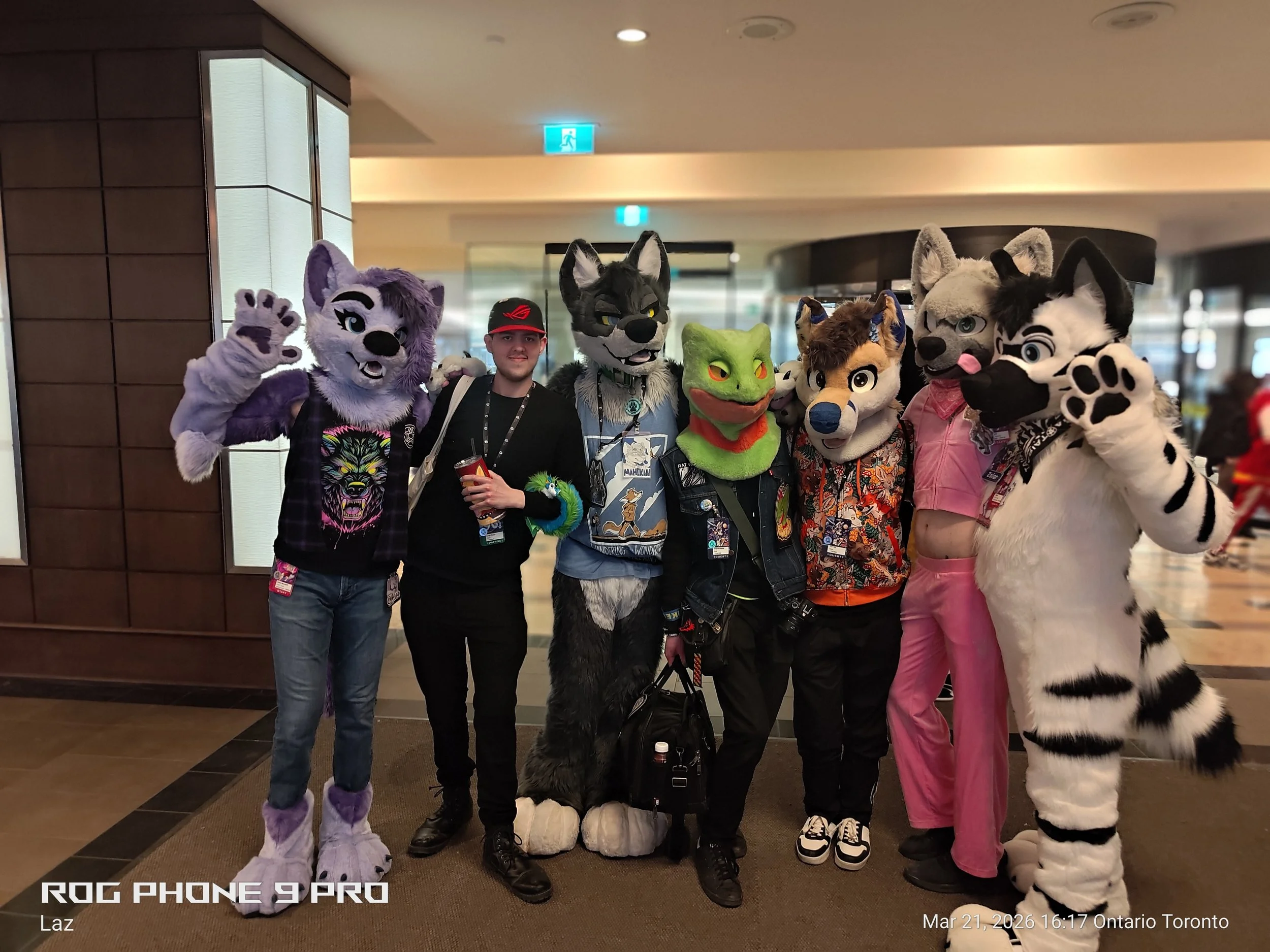 A group of people dressed in various furry animal costumes with a person standing in the middle. They are indoors at an event, with some wearing badges around their necks.