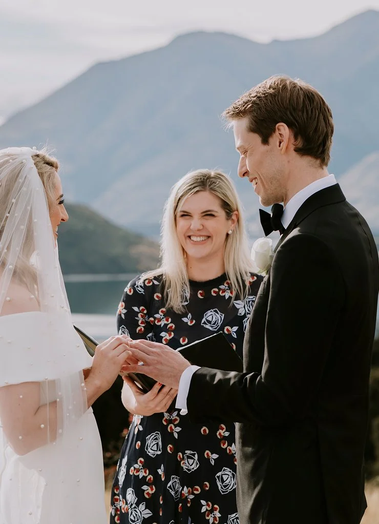 A wedding ceremony outdoors with a lake and mountains in the background. A bride and groom exchange rings, with a smiling female officiant standing between them.